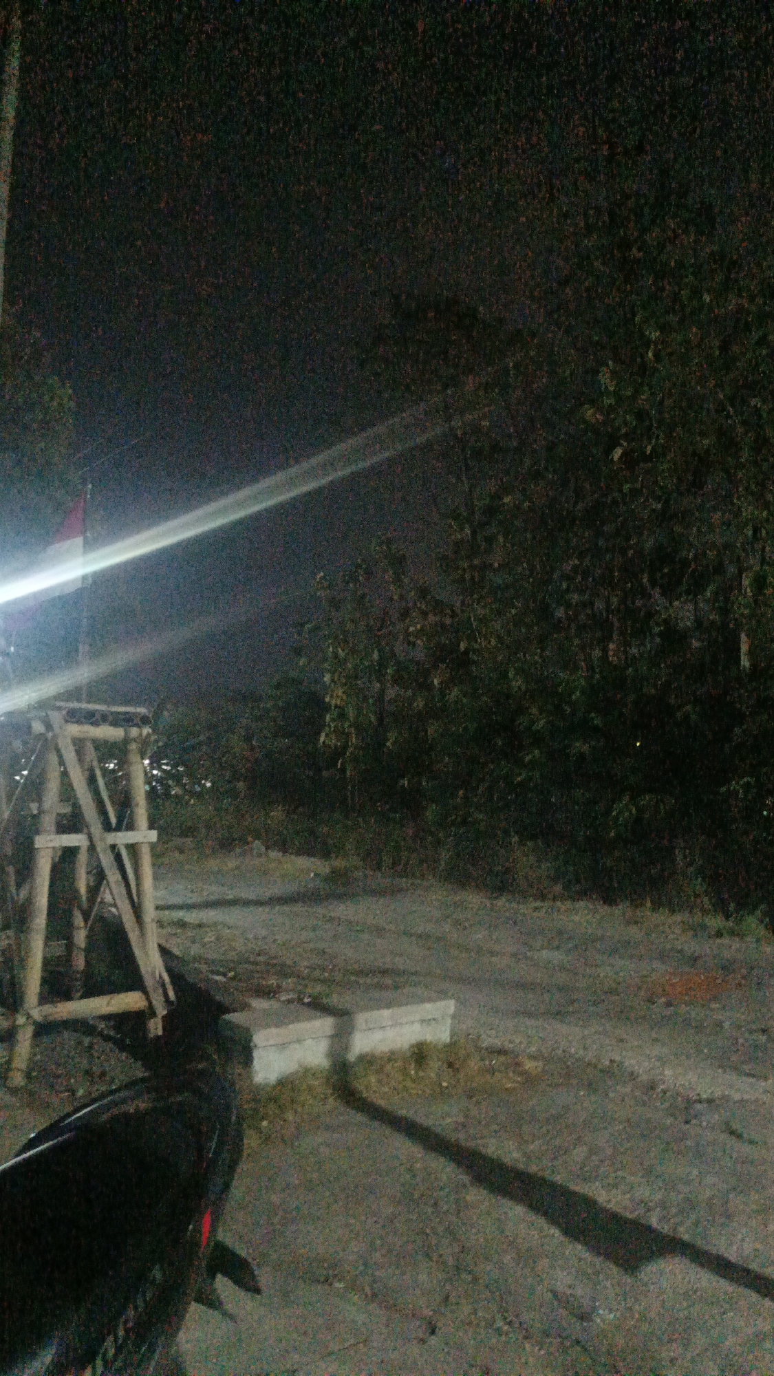 The image depicts an outdoor scene at night, likely in Sambi, Indonesia, characterized by low light and visible noise. In the foreground to the left, a portion of a dark-colored motorcycle is visible, with its rear visible and a red taillight. To the left of the motorcycle is a wooden structure that appears to be scaffolding or a temporary frame, with diagonal supports. Above and behind the wooden structure, a flag is partially visible, appearing to be red and white, consistent with the Indonesian flag. A bright light source, likely a lamp, casts a strong glare across the upper left and central portion of the image, creating a visible light streak.
The background consists of dense foliage, suggesting a natural or semi-natural environment with trees and bushes. The ground is a mix of gravel and dirt, with some patches of grass. In the mid-ground, two rectangular concrete blocks are placed on the ground, with a shadow cast diagonally across the scene from the right, indicating another light source or an object casting a shadow. The overall impression is a somewhat unkempt, dimly lit outdoor area at night.
