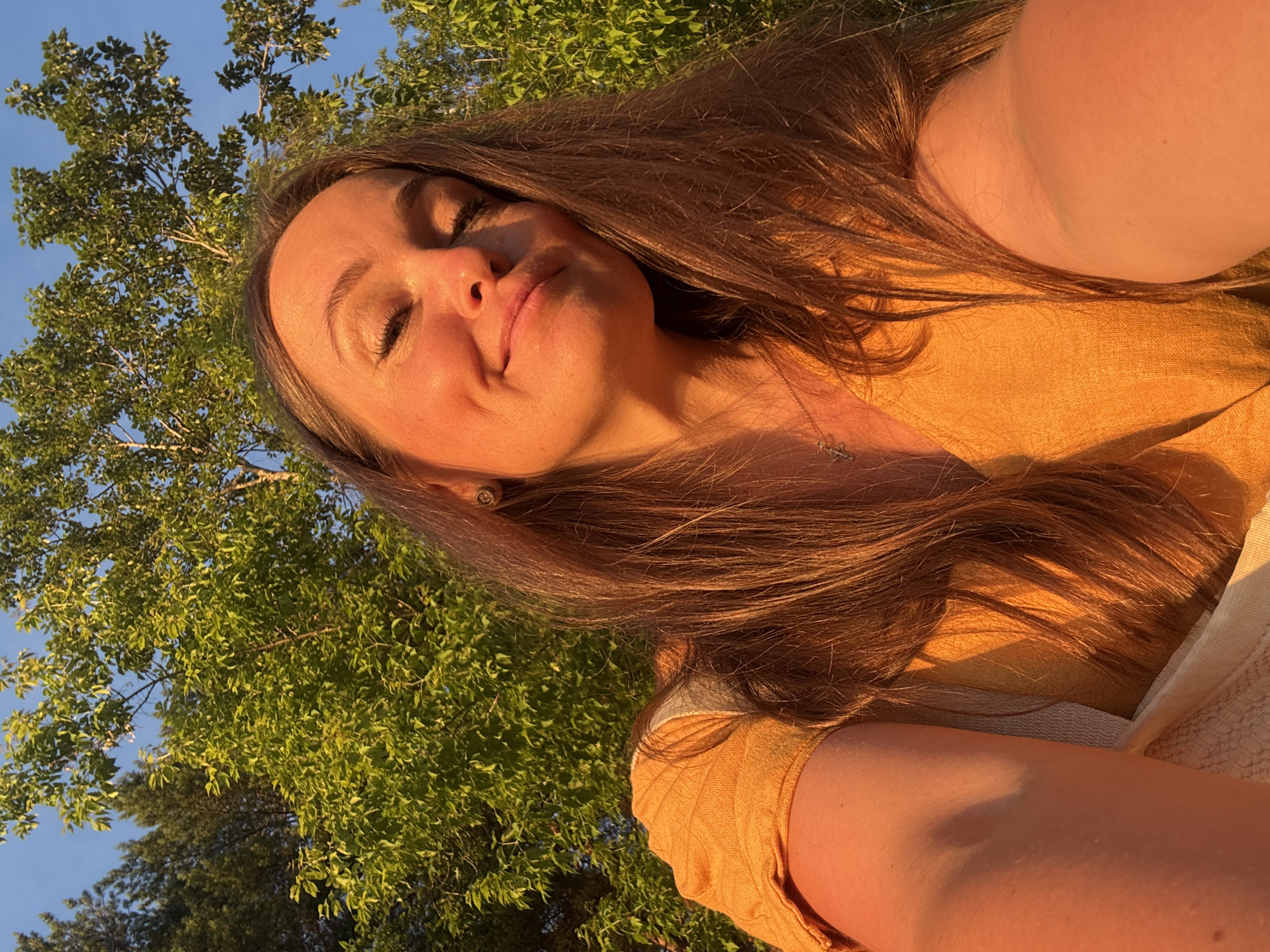 A young woman is captured in a close-up selfie, with her eyes closed and a slight smile on her face, suggesting a moment of peace or contentment. She has long, brown hair that falls around her shoulders. She is wearing a yellow top, and a faint glimmer of a necklace with a cross pendant is visible. The lighting indicates it is either sunrise or sunset, casting a warm, golden glow on her skin and hair.
The background features lush green foliage and a clear blue sky, suggesting an outdoor setting in a natural environment. The trees are dense and vibrant, implying it's a warm season, likely summer. The orientation of the photo, taken from a low angle looking upwards, further emphasizes the natural surroundings. The scene evokes a feeling of relaxation and appreciation for nature. The image does not contain any visible text. Given the location context of Chaikovsky, Russia, the setting is likely a park, garden, or a natural landscape near the city.