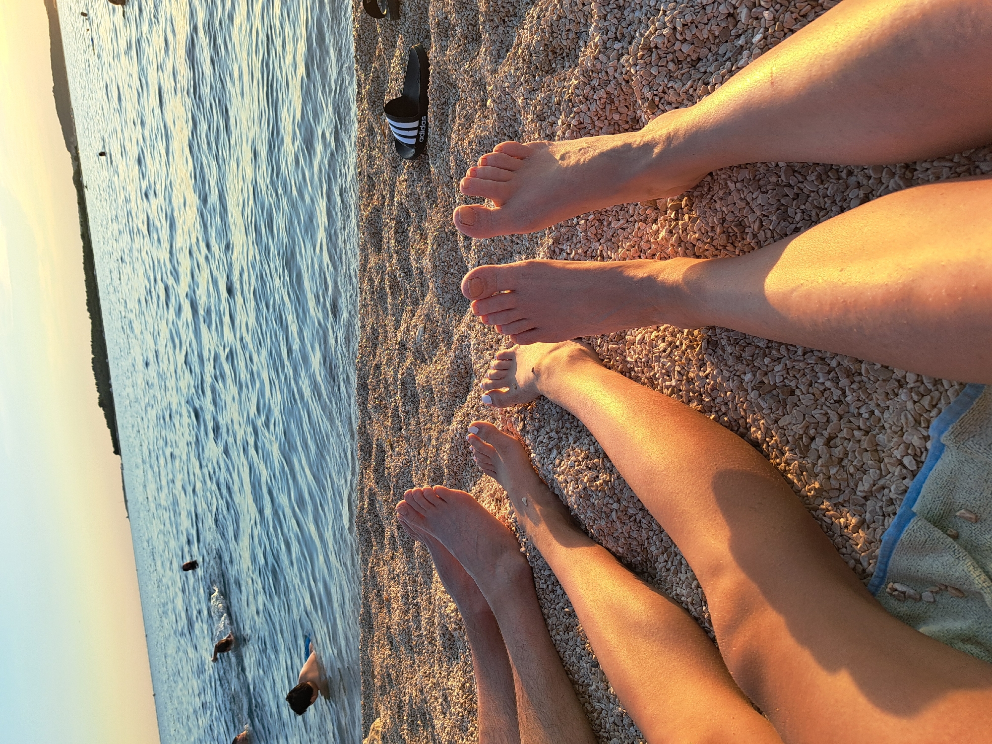 The image shows a close-up, overhead view of several people's legs and feet resting on a pebble beach next to calm water. The legs and feet are positioned diagonally across the frame, with some in the foreground and others further back. The pebbles are a mix of light brown and grey, and the water on the left is a deep blue with gentle ripples, reflecting the warm light of the setting sun. A black Adidas sandal with white stripes is visible on the pebbles near the top left of the image. The lighting suggests it is late afternoon or early evening, with a warm, golden glow illuminating the scene. The overall mood is relaxed and peaceful, evoking a sense of a summer day at the beach. There is also a suggestion of people swimming or wading in the water in the background, though they are out of focus.
