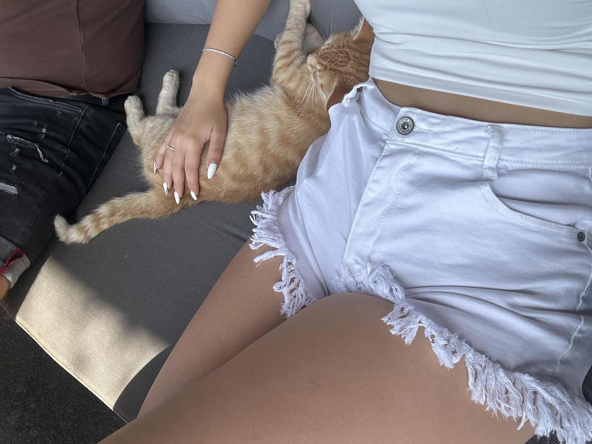 In Bania, Bulgaria, a person is lounging on a sofa outdoors, with a ginger cat resting on their lap. The person is wearing white denim shorts with frayed edges and a white top. The cat is lying on its back, with its paws extended. Sunlight casts a shadow across the scene, creating a high-contrast environment.