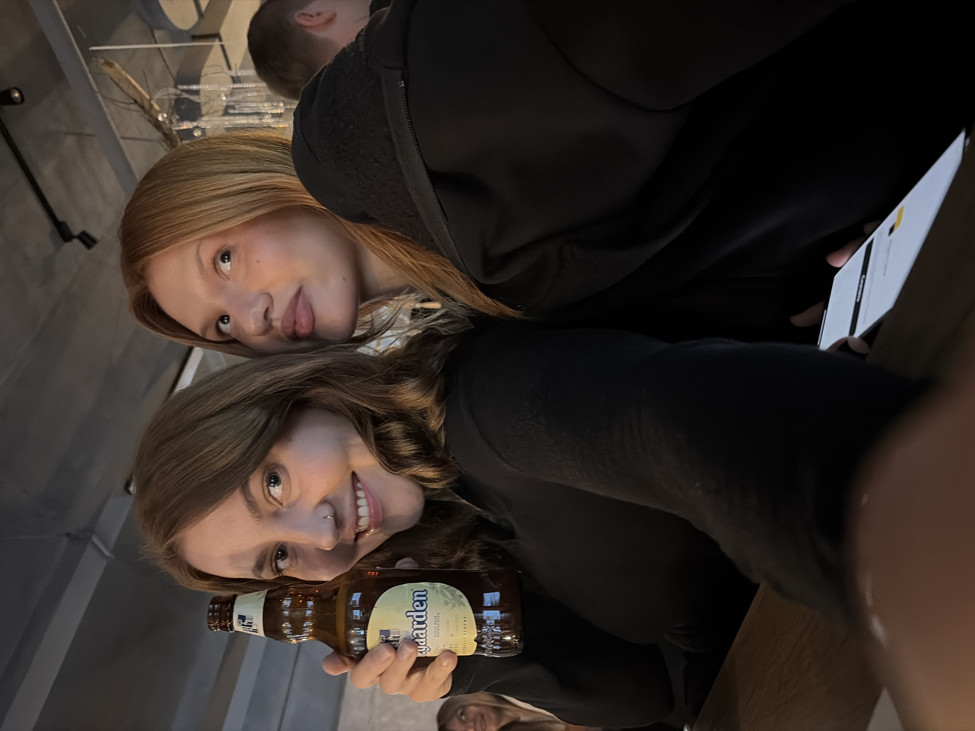 Two young women are pictured, likely in a bar or restaurant setting in Chaikovsky, Russia. The woman on the left is holding a bottle of beer, possibly a "Lykkegaard" brand, and smiling at the camera. She has brown hair and is wearing a black top. The woman on the right, with reddish-blonde hair, is looking away from the camera with a playful expression, her tongue slightly out. She is wearing a black hooded jacket. The background is dimly lit and shows elements of a ceiling with exposed beams and industrial lighting, suggesting a casual, modern interior. Another person is partially visible in the background to the right. The overall mood appears relaxed and social.