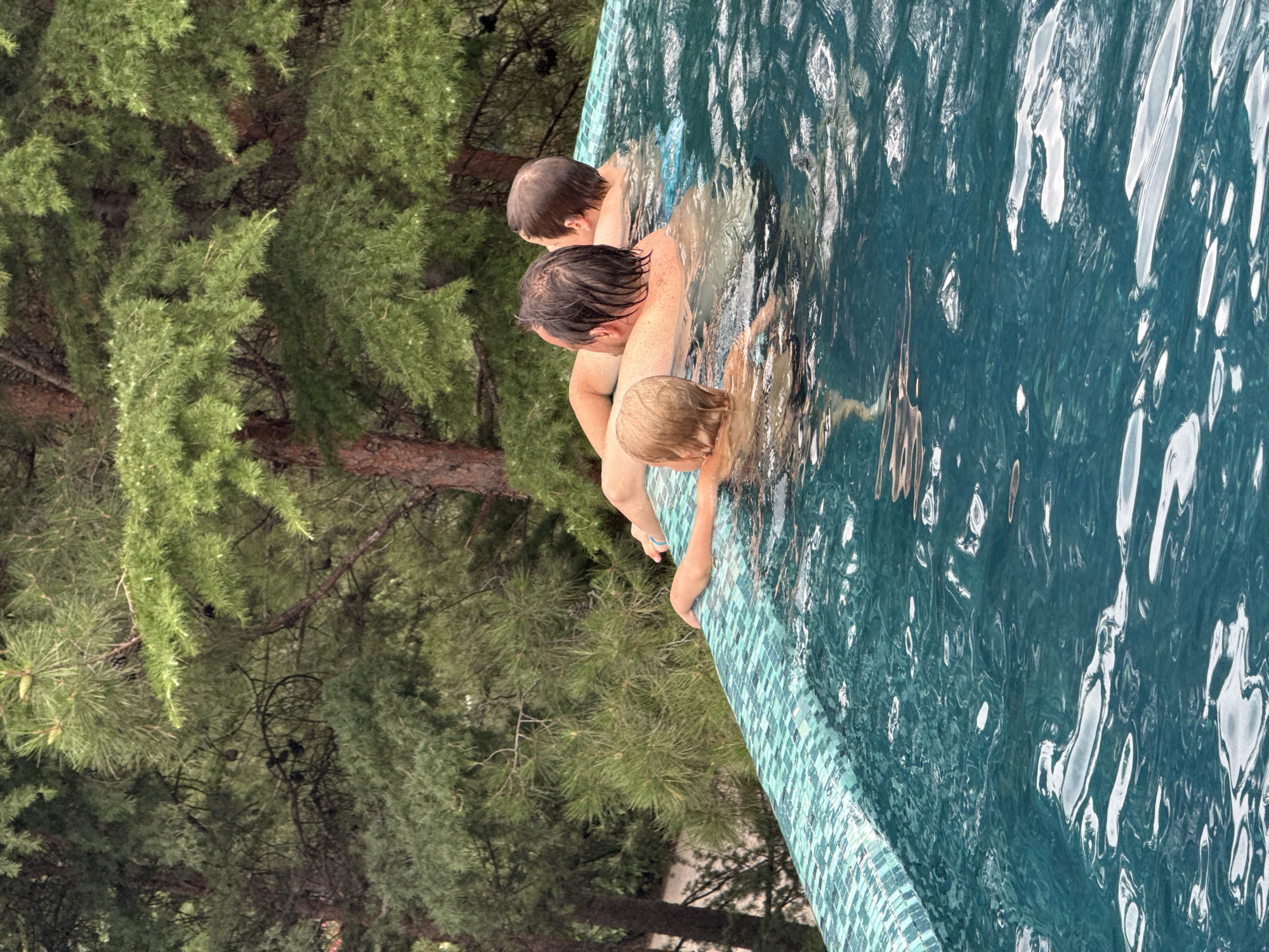 The image shows three people in a swimming pool. The pool has a mosaic tile border. The background consists of lush green trees, suggesting an outdoor setting. Two adults and one child are visible in the water, appearing to be relaxed and enjoying themselves. The lighting suggests it is daytime. The location is not specified within the city of Yalta, Ukraine, and the provided context does not offer any information about this.