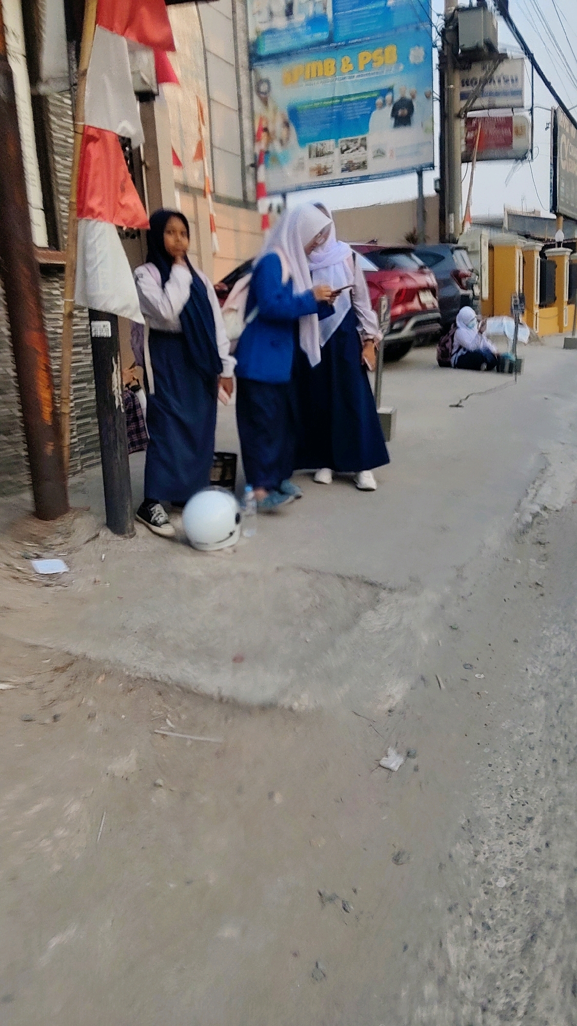 A group of three young females, dressed in school uniforms consisting of long skirts, long-sleeved shirts, and headscarves, are standing on a paved street in front of a building with a prominent billboard in Mekarmukti, Indonesia. The female on the left, wearing a navy hijab and a white shirt, is looking to her right. The middle female, with a light purple hijab and a blue jacket, is holding a mobile phone and looking at it. The third female, also with a light purple hijab, stands to her right, partially obscured. In the foreground, a white helmet rests on the ground near the feet of the middle and left females, alongside a plastic water bottle. A red car is parked behind them, and another person, also in a school uniform, is seated on the ground further down the street. The environment appears to be an urban street with some debris on the ground, including paper scraps and small sticks. The lighting suggests it is either early morning or late afternoon.
