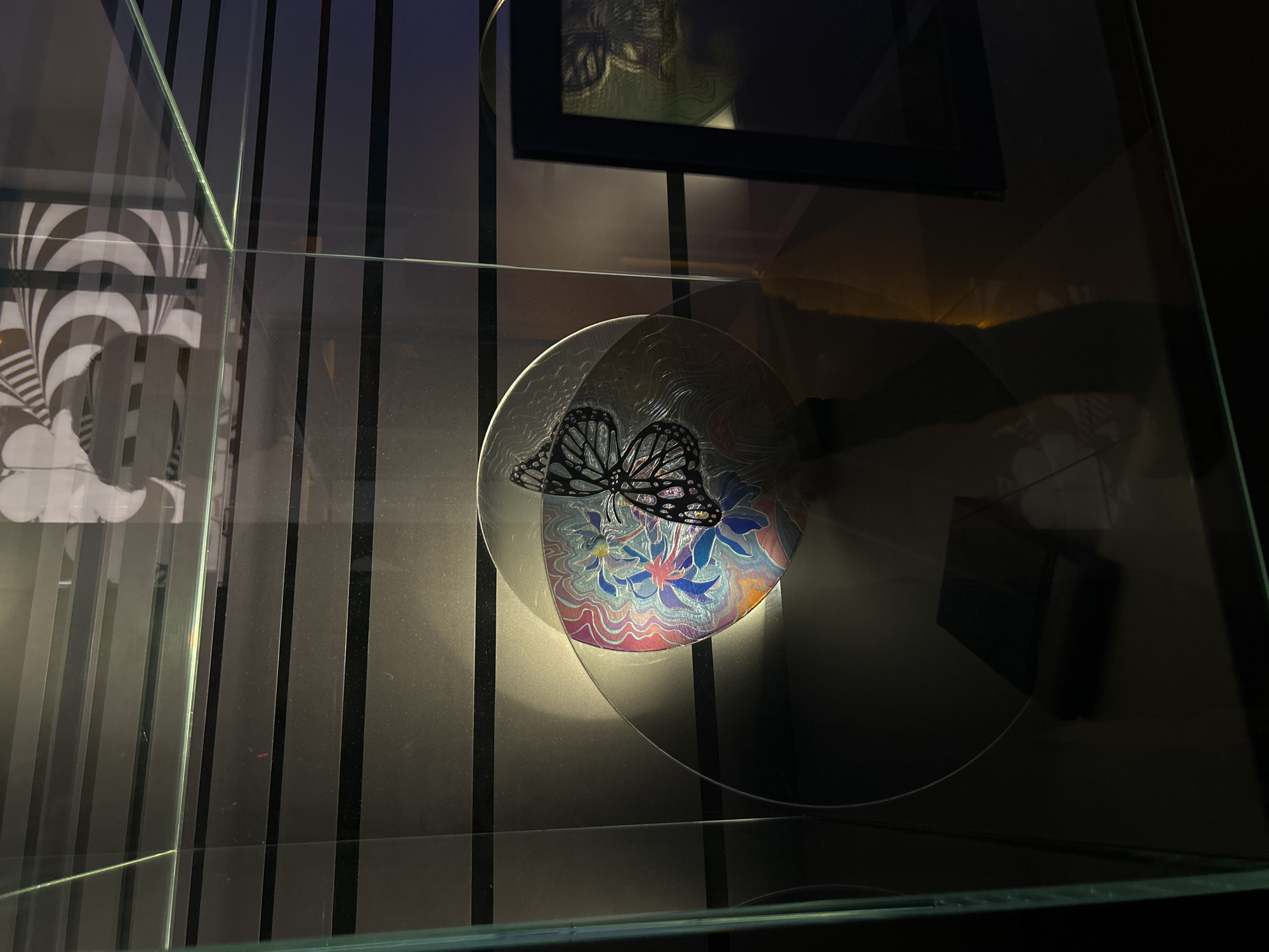 The image shows a display case, likely in a museum or gallery, containing several art pieces. The background features vertical lines of varying shades of gray and black. In the foreground, a clear, round disc with a butterfly design on it is prominently displayed. The butterfly's wings are black with intricate patterns. The disc also has floral and wave patterns in shades of blue, pink, and white. Above the disc is a black-framed artwork, though only a portion of it is visible. The display case is made of glass and has reflective surfaces. The location of the image is Unknown, Unknown.