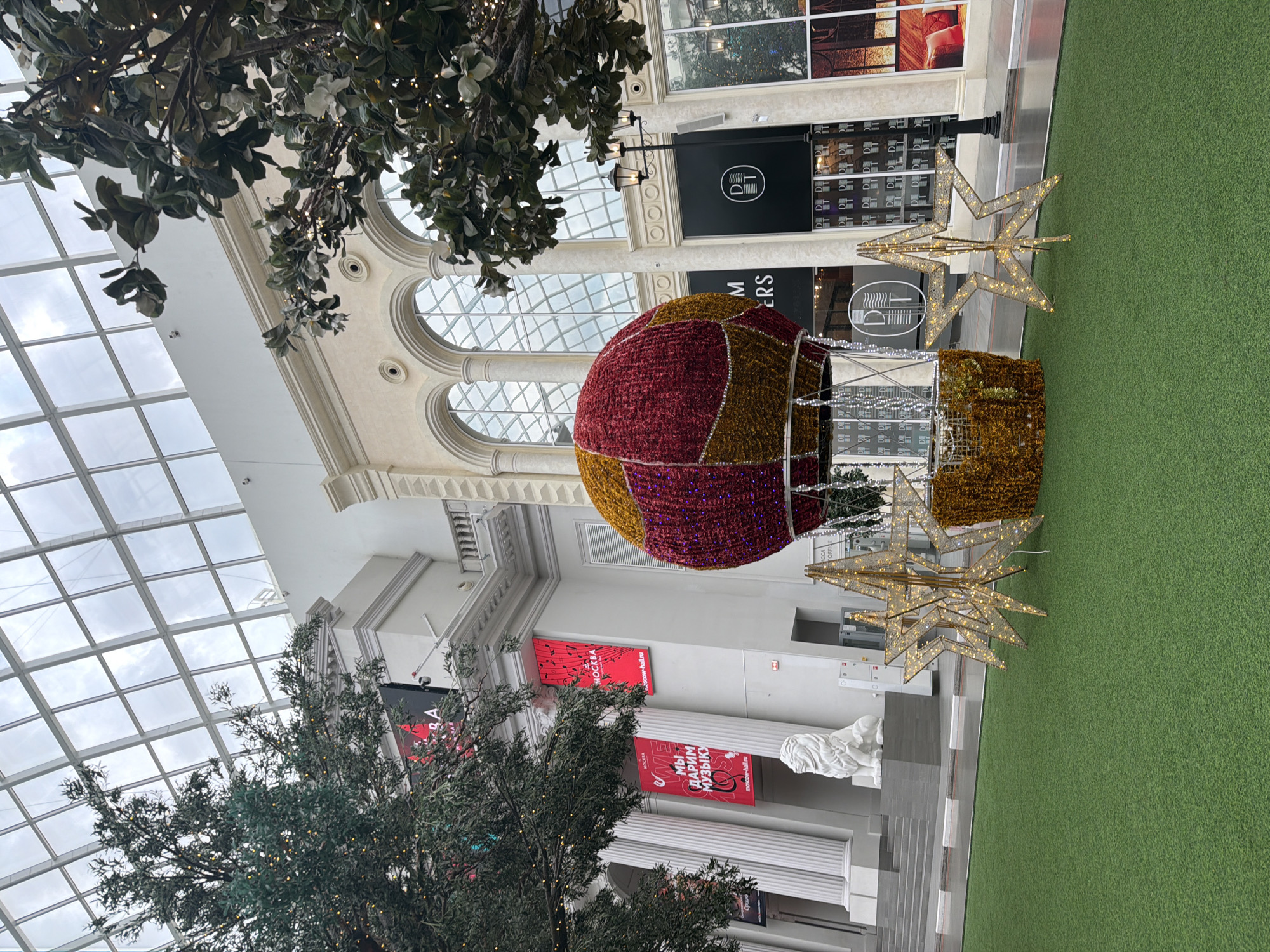 This is a vertical, eye-level, wide shot of a festive indoor scene, possibly a shopping mall or a public atrium decorated for the holidays. The lighting suggests daytime, with natural light filtering through a large glass ceiling. The overall mood is cheerful and decorative.
The main subjects are several large, illuminated decorations. A prominent hot air balloon, constructed from red and gold sparkling material, hangs in the center. Next to it are several large, star-shaped lights, also illuminated. A tall, artificial tree adorned with small, twinkling lights is visible in the foreground on the left.
The setting is an indoor space with classical architectural elements, including large white columns and arched windows. The floor is covered with artificial green turf on the right side of the frame. In the background, there are storefronts with displays.
Several pieces of text are visible:
- A black sign with a white circle and stylized logo reads "DT" inside the circle.
- Another black sign reads "DM" and "ERS".
- A red banner with musical notes and the text "МОСКВА" (Moscow) is visible.
- Another red banner has Cyrillic text that translates to "WE GIVE MUSIC."
- A white bust of a sculpted head is placed on a pedestal near the columns.
There are no people visible in the frame. The scene appears to be set up for a holiday event or promotion.