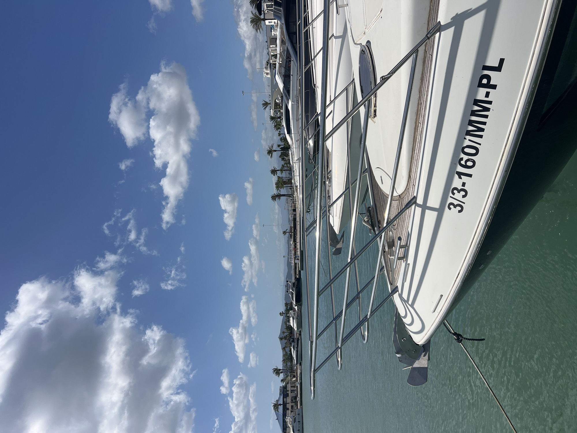 A daytime scene presents a yacht berthed in M'Diq, Morocco. The yacht, predominantly white, is moored with a rope visible near its bow. Its hull displays the inscription "3/3-160/MM-PL." A chrome railing runs along the side of the yacht.
In the background, palm trees line the waterfront, punctuated by buildings. Other yachts are docked nearby. The sky is blue with scattered cumulus clouds. The water is calm and greenish-blue.