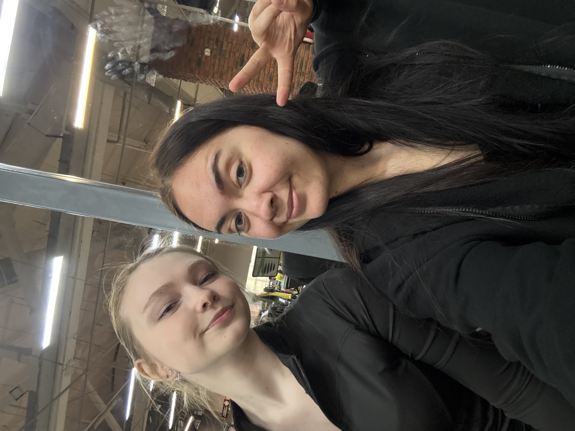 Two young women are posing for a selfie in an indoor environment. The woman on the left has light skin and blonde hair. She is wearing a black top. The woman on the right has darker skin and long, dark hair. She is wearing a black hoodie and holding up her hand in a peace sign. They appear to be in a gym or industrial-like space, based on the overhead lighting, ceiling structure and reflections in the glass or mirror in the frame, which suggests that they may be in Chaikovsky, Russia. Some exercise equipment can be seen in the background.