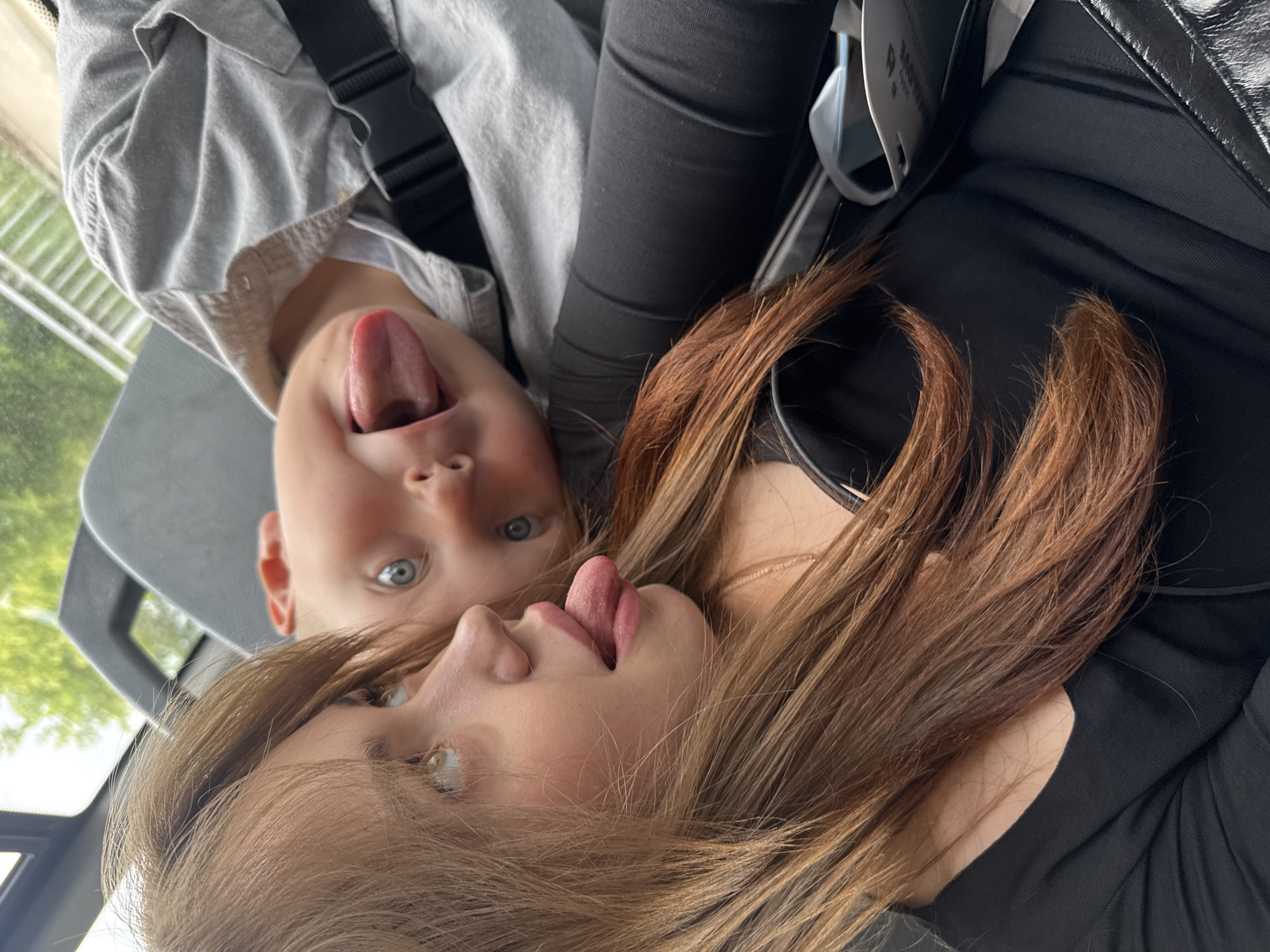 A close-up, tilted shot shows two people inside a vehicle, possibly a bus. The person in the foreground, to the left, is a young female with long, light brown hair. She is wearing a black top and is looking upwards and to the left with her mouth slightly open, her tongue sticking out. Next to her, behind and to the right, is a baby or young child. The child is lying on their back and looking upwards, sticking their tongue out with a wide grin. They are wearing a light-colored shirt. The background is blurred, showing glimpses of trees and a light-colored railing, suggesting an outdoor environment visible through a window. The overall impression is playful and candid.