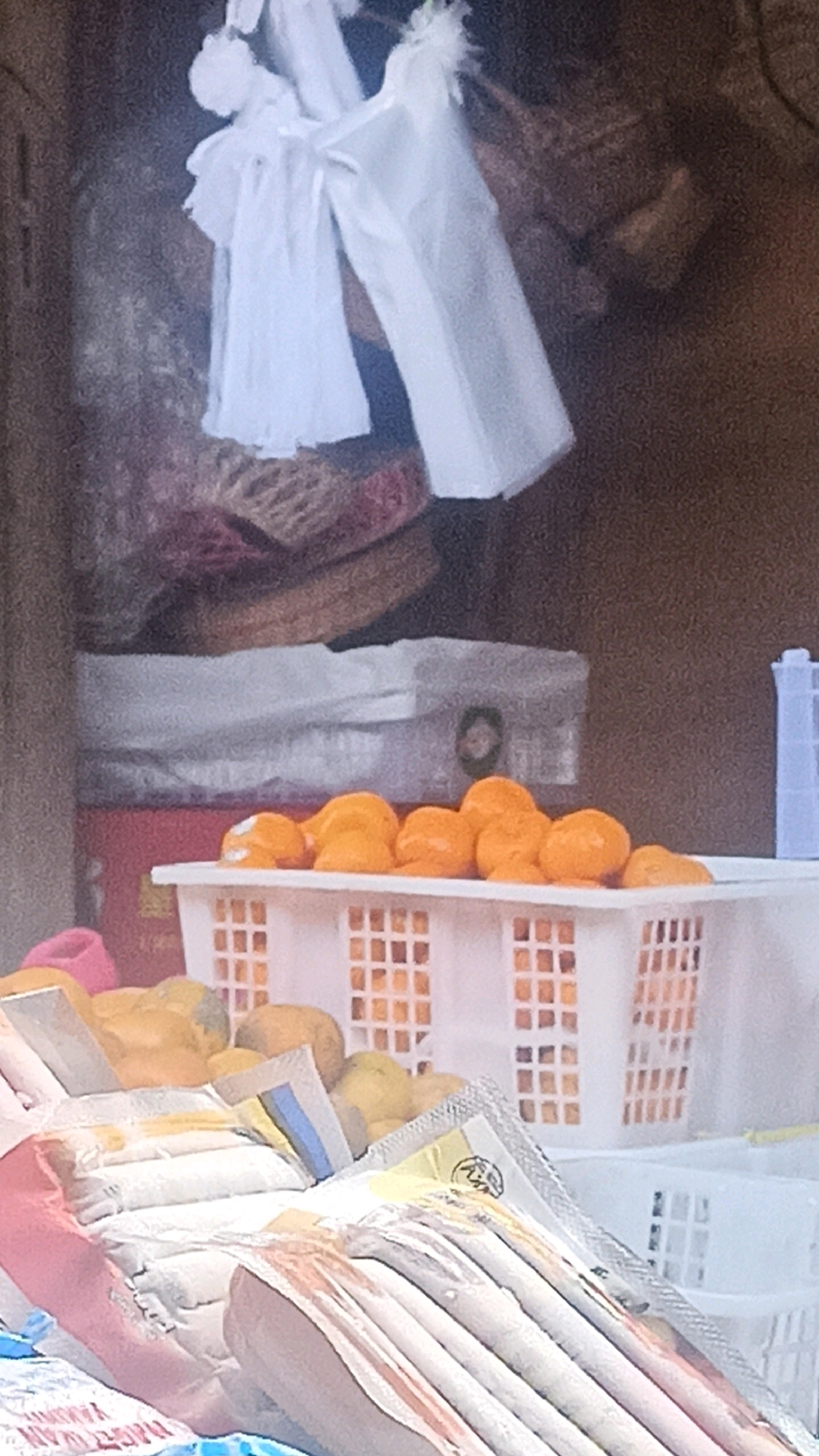 The image depicts a market stall in Tuba, Indonesia, displaying a variety of items for sale. In the foreground, a white plastic basket is filled with bright orange citrus fruits, possibly tangerines or mandarins. Behind this basket, another white plastic basket holds yellow fruits, likely papayas or similar produce. Further back, several items are visible, including what appear to be stacks of white plastic bags or packaging material. The background is darker and more ambiguous, with hints of woven baskets or mats, and some indistinct fabric textures. The overall impression is one of a bustling, perhaps outdoor, market environment.