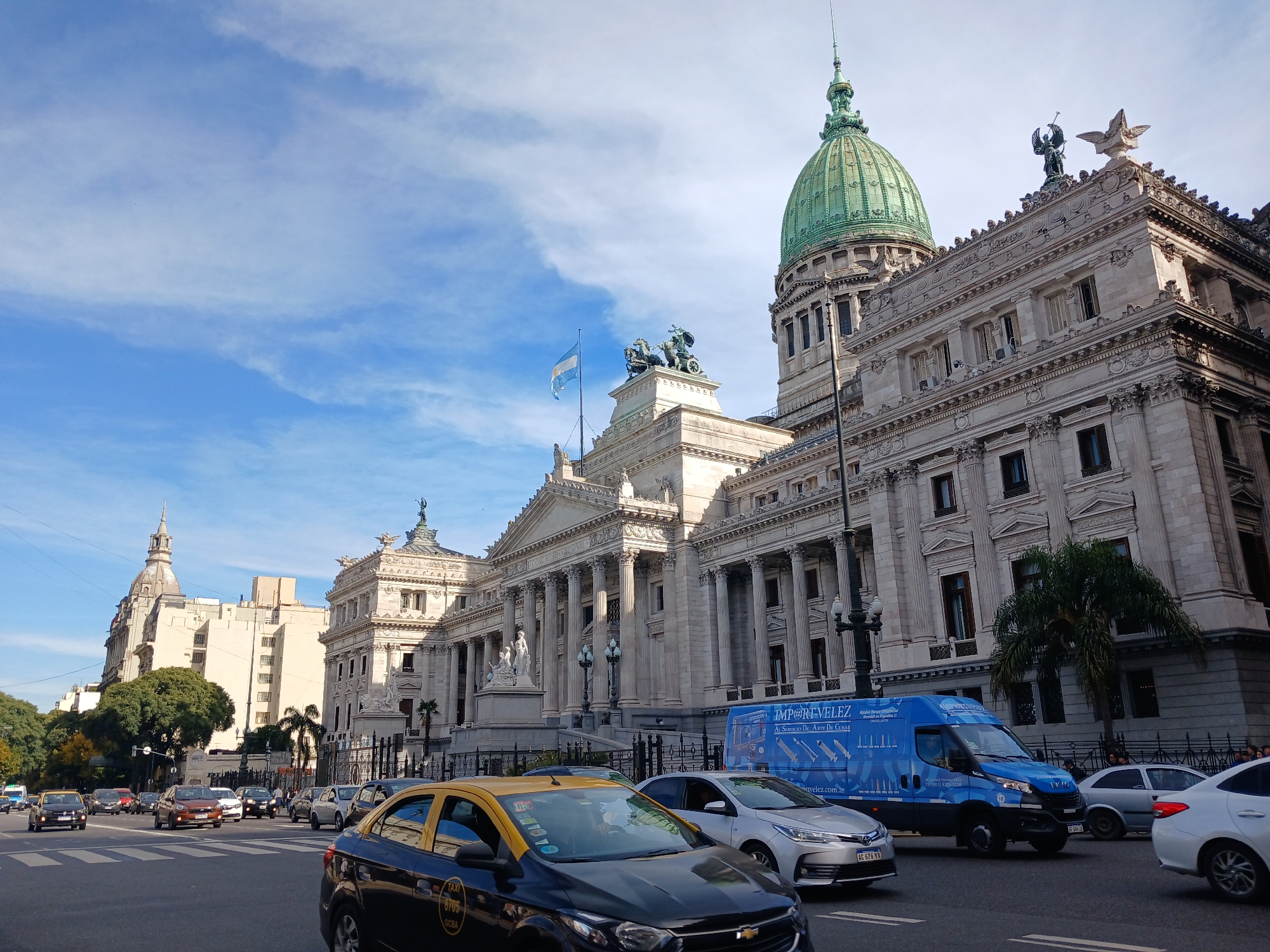 The image shows the Argentine National Congress building in Buenos Aires, Argentina, captured from a street-level perspective. A variety of vehicles, including a black and yellow taxi, a blue delivery van, and several passenger cars, are visible in the foreground. The sky is blue with scattered clouds. The building's architecture includes columns, statues, and a green dome. A palm tree is visible to the right of the frame in front of the building. An Argentine flag flies from the building's roof.