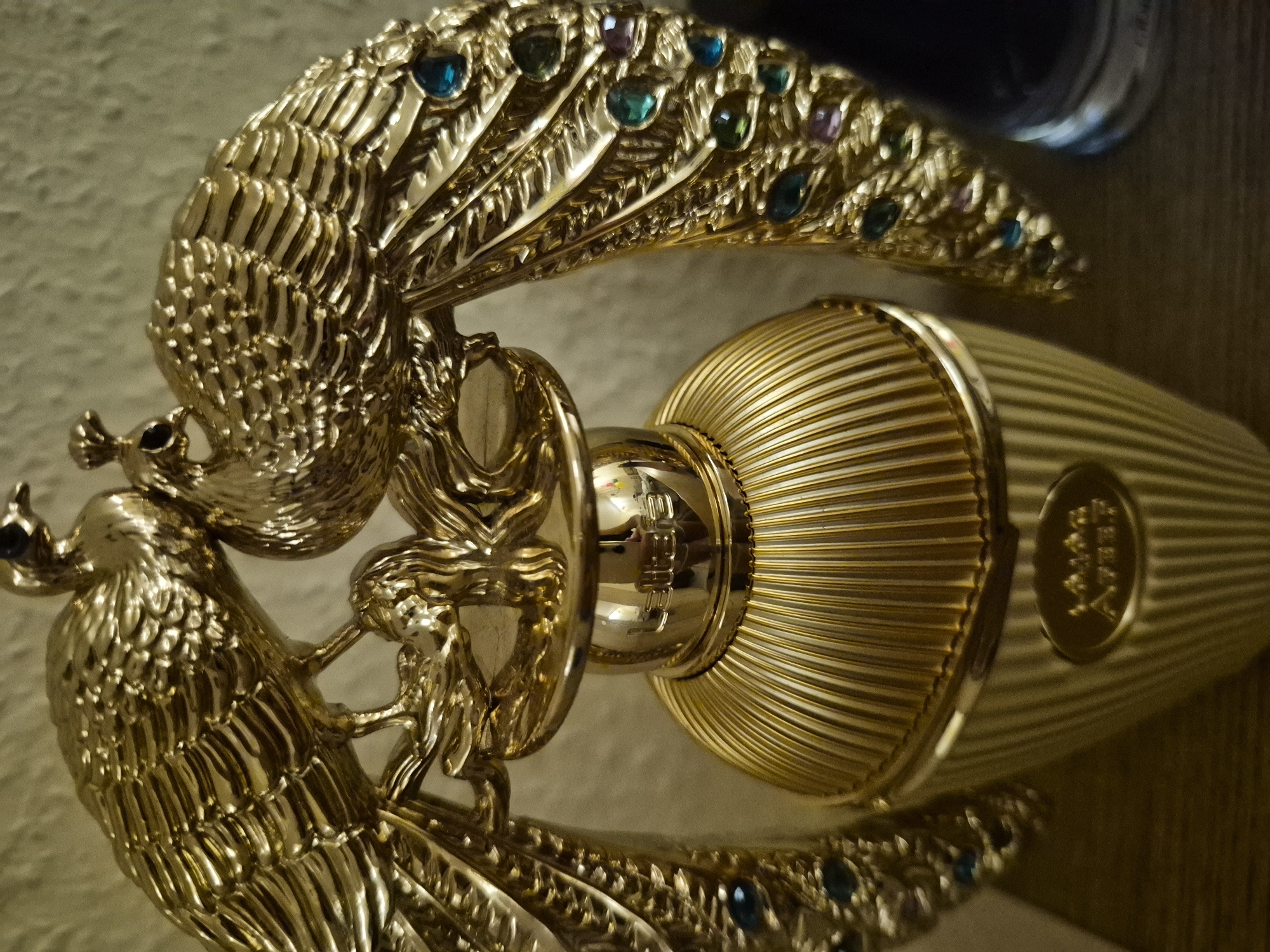 A gold-colored perfume bottle with a fluted design is prominently displayed. Two ornate gold peacocks with jeweled tail feathers are perched on the top of the bottle. The peacocks are detailed with individual feathers and appear to be intertwined. Some of the feathers are adorned with small, colorful gemstones in shades of blue, green, and purple. The bottle itself has a ribbed texture and a metallic sheen. A label is visible on the front of the bottle with embossed text, though it is partially obscured and difficult to read clearly. The background is a neutral, textured wall. The overall aesthetic suggests luxury and opulence.