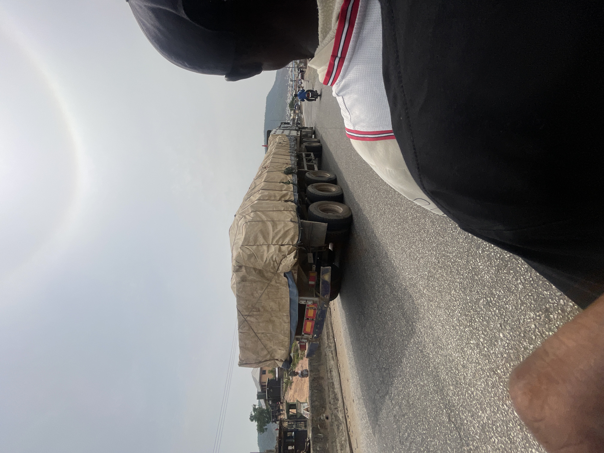 This image, taken in Lokoja, Nigeria, captures a dynamic road scene, likely from a moving vehicle. The perspective is from the right side of the road, looking forward. In the immediate foreground on the right, the head and shoulder of a dark-skinned person are visible, wearing a dark cap or hood and a white top with prominent red and blue striped detailing on the shoulder, worn over a black garment.
The central subject of the photograph is an exceptionally long, multi-axle flatbed truck, heavily loaded and completely covered by a light brown or tan tarp. The truck's numerous wheels are visible as it moves along the paved asphalt road. The road surface appears bright, possibly reflecting sunlight.
In the background, the sky is bright and overcast, revealing a striking solar halo – a large, faint rainbow-like ring encircling the sun, which is obscured by the clouds. Beyond the truck and road, the environment features various buildings, some greenery, and distant hills or mountains, characteristic of a semi-urban landscape. Other vehicles, including a person on a motorcycle, and pedestrians can be seen further down the road, indicating active traffic. The time of day appears to be bright, likely midday or afternoon, given the intensity of light and the solar halo. There is no discernible text visible in the image.