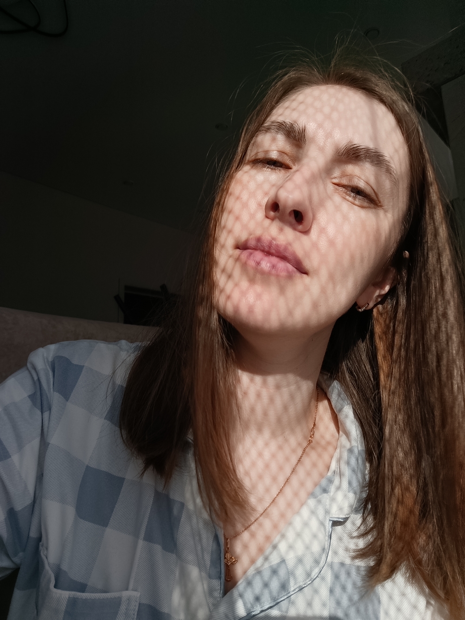 A woman with light brown hair is facing the camera. She is wearing a blue and white checkered shirt and a gold necklace with a cross pendant. Her eyes are partially closed, and her face is covered in a patterned shadow. The background is dark, with a glimpse of a beige surface. The location is unspecified and cannot be determined from the image.