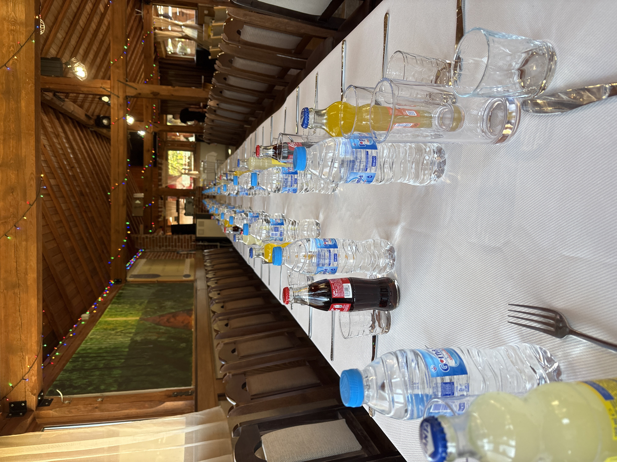 The image shows a long table set for a meal, likely a formal or celebratory occasion. The location is probably indoors, given the wooden beams and decorative lights in the background, and the description states it's in Kopilovtsi, Bulgaria.
The table is covered with a white tablecloth. There are chairs lined up on both sides, suggesting a banquet-style arrangement. Each place setting consists of a water bottle, a small bottle of juice or soda, a glass, and silverware (a fork and knife). The placement suggests that the table is set for a large group of people. The focus of the image is on the long, prepared table rather than on any specific individuals or activities.
