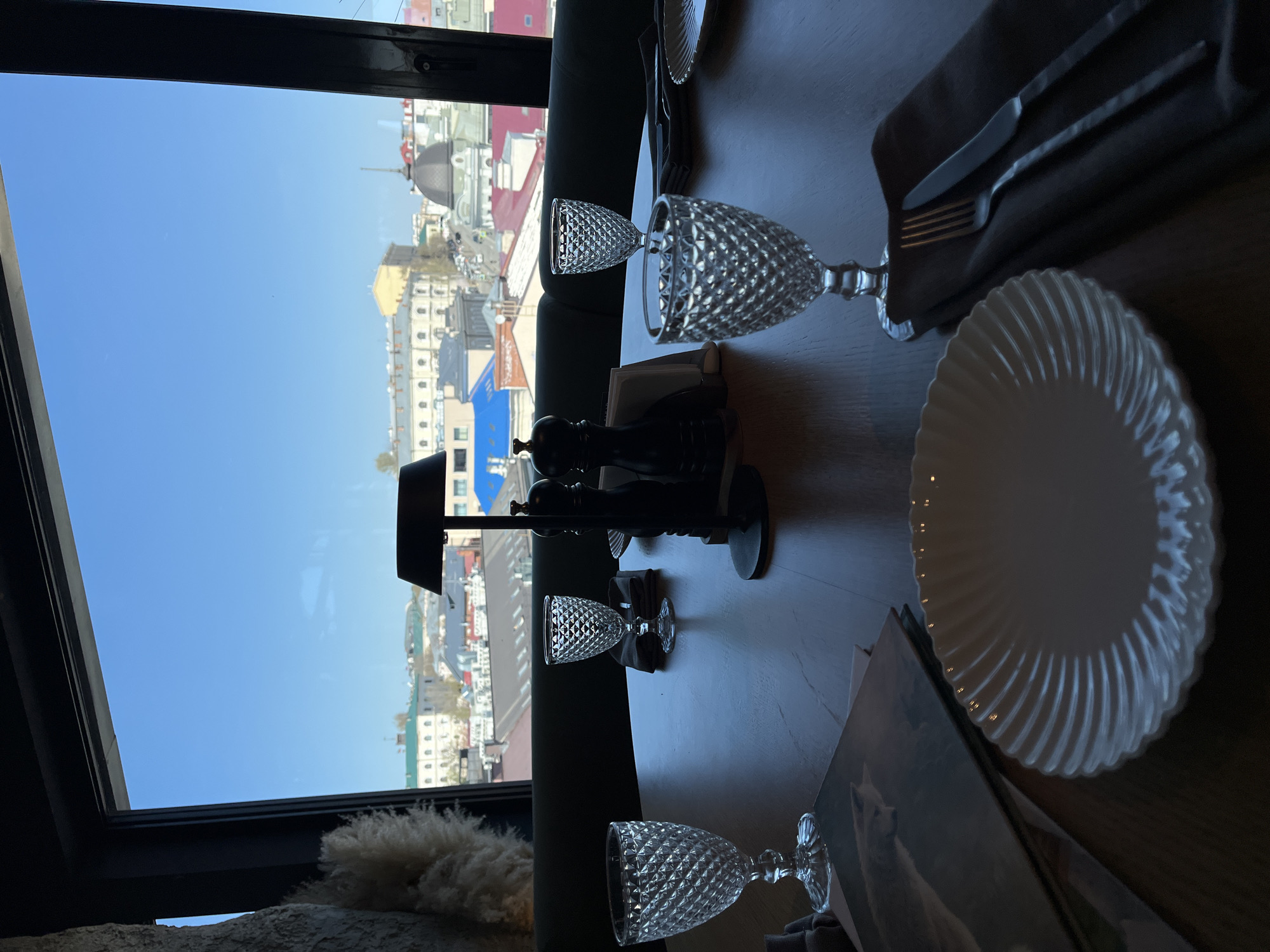 The image shows a table setting in a restaurant in Kazan, Russia. A window offers a view of the city. The table is set with decorative clear stemware glasses, white plates with a ridged pattern, silverware wrapped in gray napkins, a small lamp, and pepper mills. There is a book on the table. The day appears sunny.