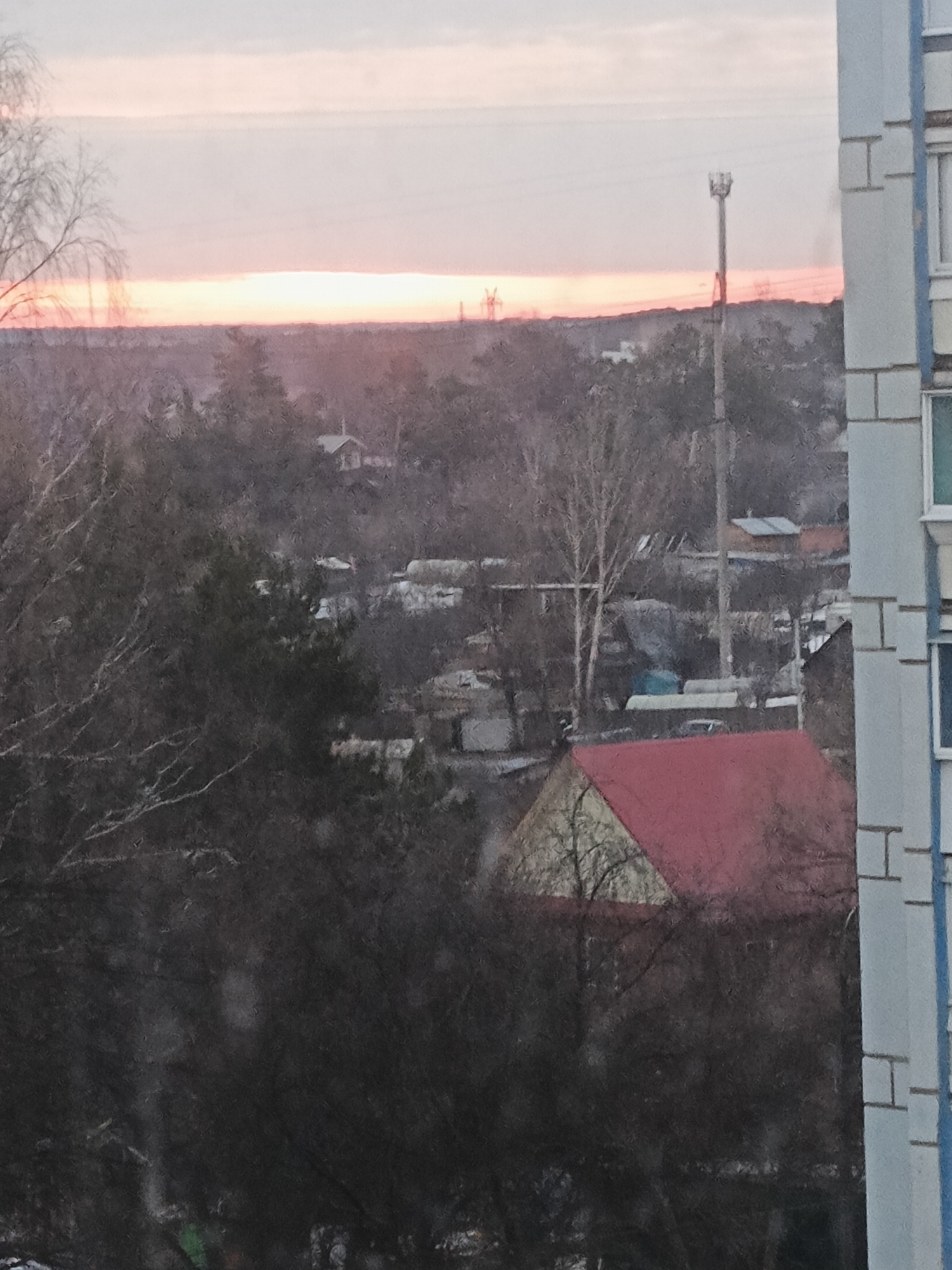 The photograph captures a cityscape during a sunset or sunrise, taken from a window in a building located in Chaikovsky, Russia. The sky exhibits hues of pink and orange along the horizon. A red roof is partially visible in the midground, obscured by bare trees. Behind the trees, a collection of houses and smaller buildings are nestled within a wooded area. In the background, utility poles and transmission towers are silhouetted against the sky. The edge of a multi-story building is visible on the right, featuring rectangular architectural elements.