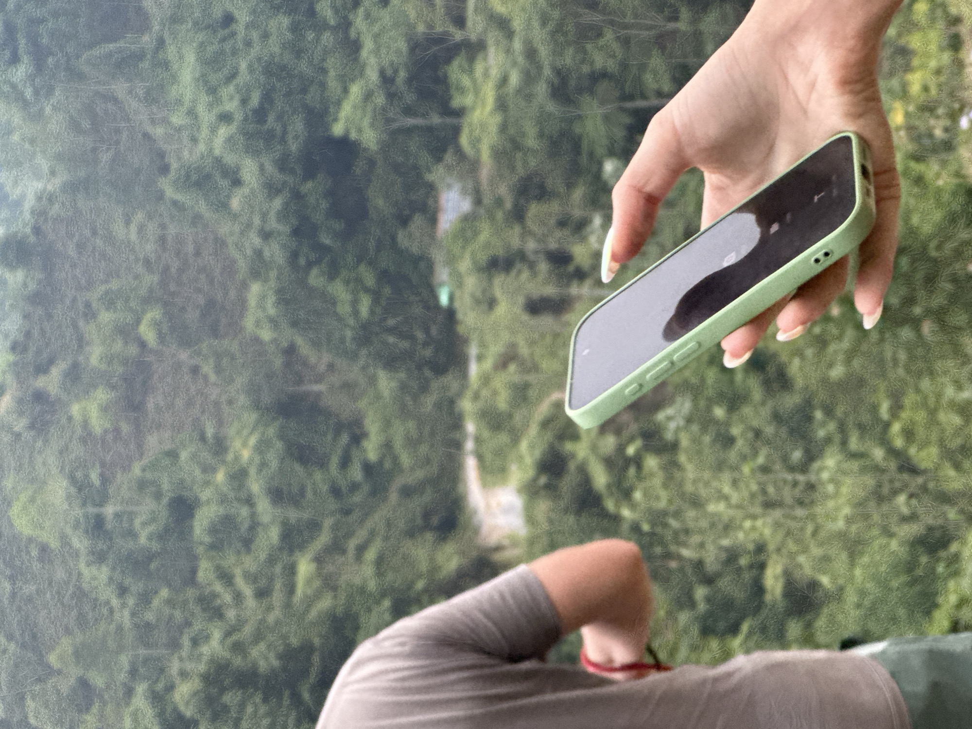 The image is presented in a vertical orientation, showcasing a person's hand holding a smartphone against a background of a dense, green forest. The primary subject is a light-skinned hand, likely female given the shape and painted fingernails, positioned on the right side of the frame. This hand holds a smartphone with a light green case, its dark, reflective screen angled slightly downwards.
In the lower left quadrant, a portion of another person's body is visible, including a shoulder and arm clad in a grey garment. A red band or bracelet adorns the wrist of this arm. Both individuals appear to be looking out or down at the scenery.
The background depicts a vast expanse of lush, green forest, suggesting a high vantage point, possibly from an elevated structure or a natural overlook. Through the canopy, a winding light-colored path or narrow road can be faintly seen cutting through the trees. The light conditions indicate it is daytime, with clear visibility. The setting strongly suggests a natural, forested area, consistent with the location context of Nậm Dịch Commune, Vietnam. No visible text is present.