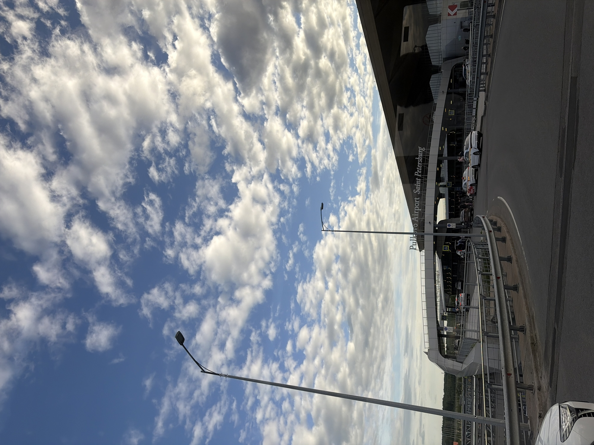 The image depicts Pulkovo Airport in Saint Petersburg, Russia, under a sky filled with numerous white clouds against a blue backdrop. The airport building is visible, along with a road, guardrails, streetlights, and several parked cars. People appear to be present in front of the airport terminal.