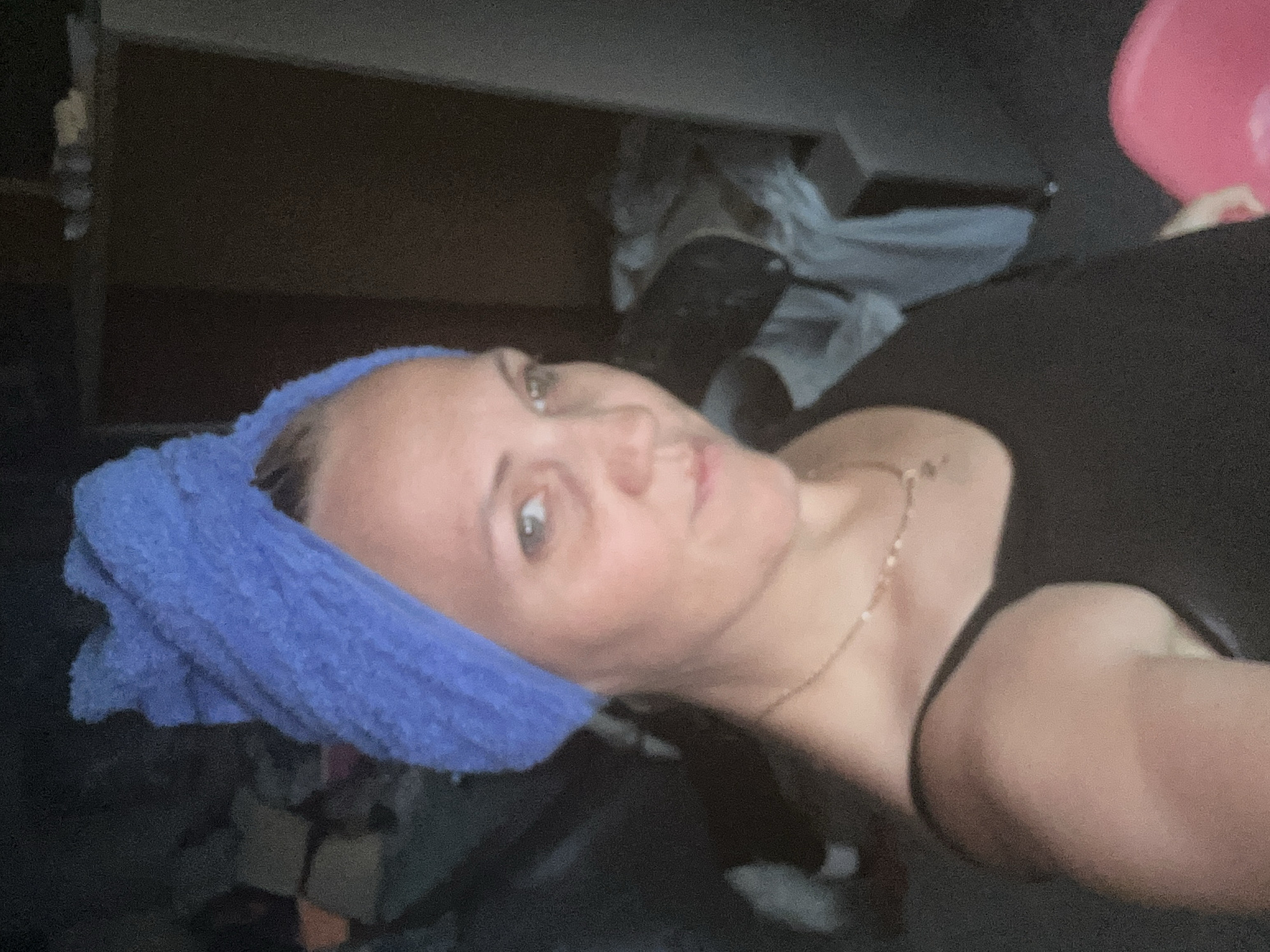 The image shows a close-up, waist-up shot of a woman with a blue towel wrapped around her head. She is wearing a black tank top and a delicate gold necklace. Her expression is neutral, with her gaze directed slightly off-camera. The background appears to be an indoor space, possibly a cluttered room with indistinct objects and furniture. The lighting is dim, casting shadows and obscuring some details of the environment. There is no indication of the specific location in Gelendzhik, Russia, nor any actions or interactions to describe.