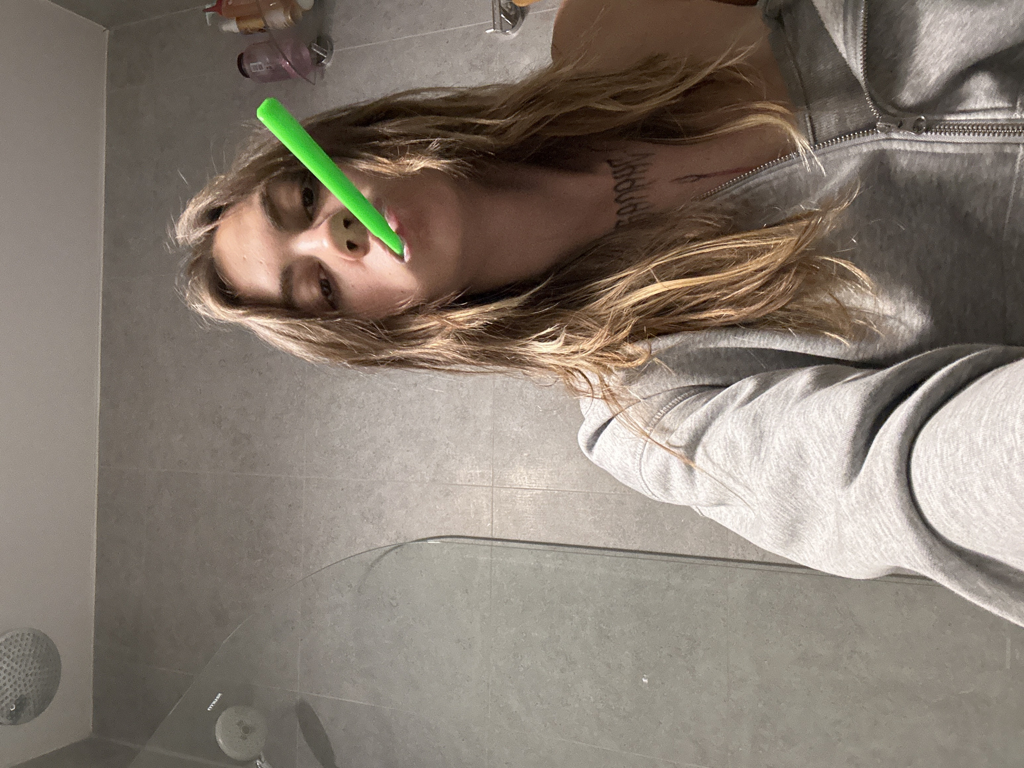 The image is a close-up selfie taken in a bathroom. A young person with shoulder-length, light brown hair is holding a bright green object, possibly a toothbrush or a pen, in their mouth. They are wearing a grey zippered hoodie. The background shows grey tiled walls, a shower head, and some bottles on a shelf. There are no discernible landmarks or context to place the image in Moscow, Russia.