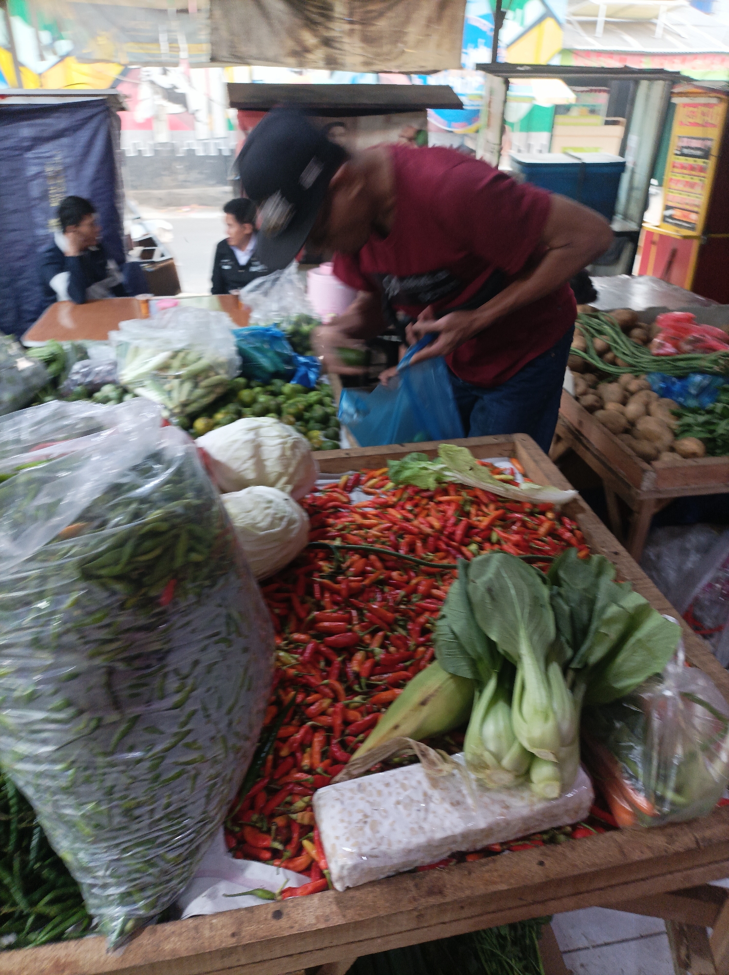 The image depicts a busy outdoor market scene in West Jakarta, Indonesia. In the foreground, a wooden stall is laden with fresh produce. Prominently displayed are large plastic bags filled with green chilies and a pile of red chilies. Two cabbages and some bok choy are also visible. Further back, a man wearing a red t-shirt and a black baseball cap is interacting with a blue plastic bag, possibly making a purchase or bagging items. Two other individuals are partially visible in the background, one wearing a dark jacket and the other a white shirt, suggesting a bustling commercial environment. The overall setting appears to be a typical Indonesian market with various goods for sale, under a makeshift roof or awning.