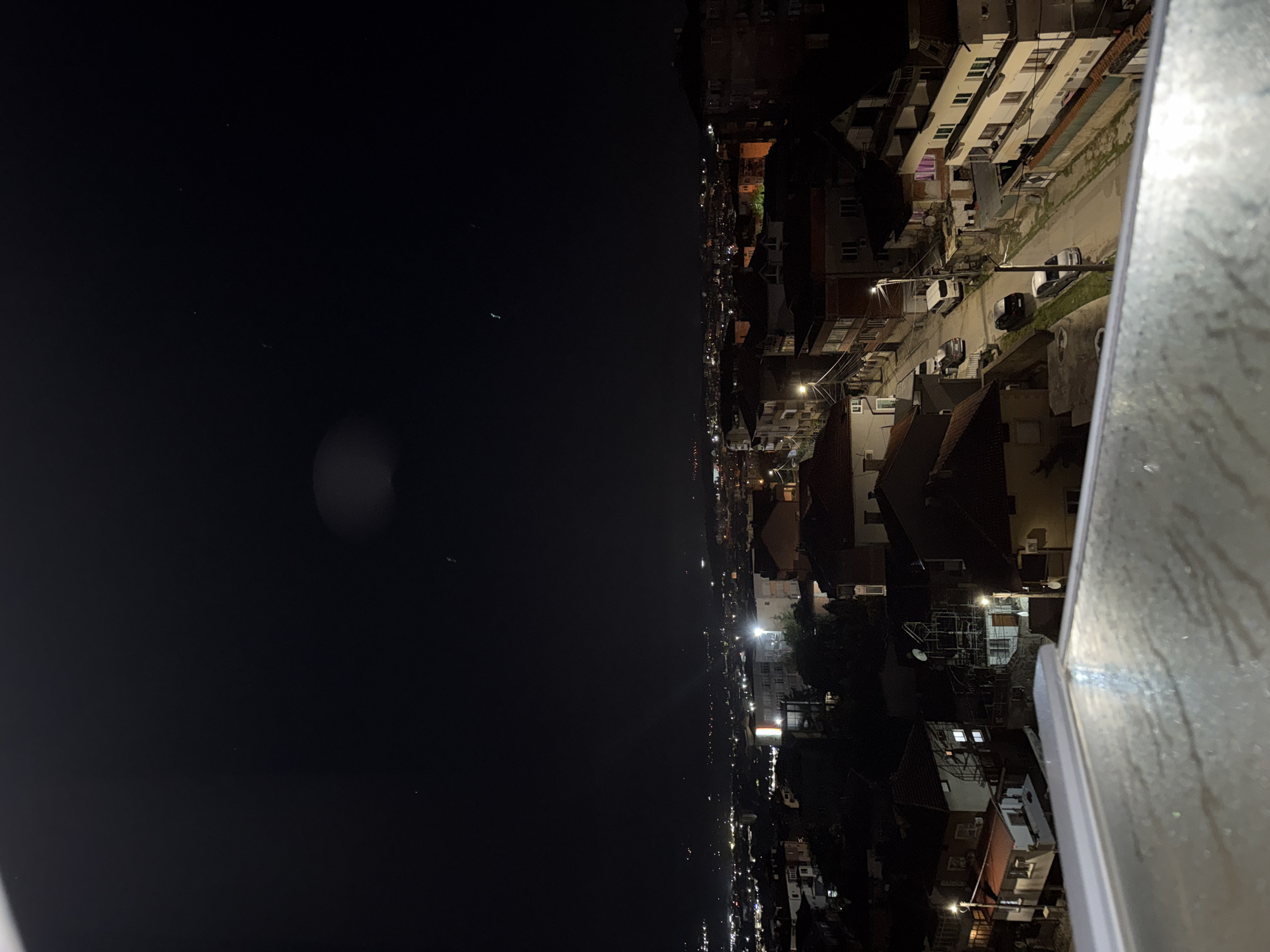 The media file presents a panoramic nighttime view of Blagoevgrad, Bulgaria, captured from an elevated perspective, likely a balcony or rooftop. The scene is dominated by a vast, dark night sky in the upper portion, exhibiting a deep black hue with a few faint, scattered points of light that could be stars or distant atmospheric phenomena. A soft, circular blur of light, possibly the moon or a distant light source, is visible in the upper-left area of the sky.
Below the expanse of the sky, the cityscape stretches horizontally across the middle and lower parts of the frame, indicating a dense urban settlement. Numerous residential and commercial buildings are visible, illuminated by a mix of warm orange-yellow streetlights and brighter white internal building lights. These lights create distinct pockets of illumination, highlighting architectural details like windows, balconies, and roofs. The buildings appear multi-story, with some exhibiting typical European architectural styles.
A street runs through the urban fabric, with several parked cars visible along its edges, illuminated by the streetlights. Utility poles and overhead wires are also discernible. On the far right, a metallic railing, possibly from a balcony, is prominently featured in the foreground, showing some texture and reflection.
The time of day is clearly night, with no natural daylight. The weather appears clear, with no visible clouds, rain, or fog, allowing for a relatively unobstructed view of the urban lights. There is no specific activity or event taking place, but the illuminated city implies ongoing life and habitation. No discernible text is visible in the image.