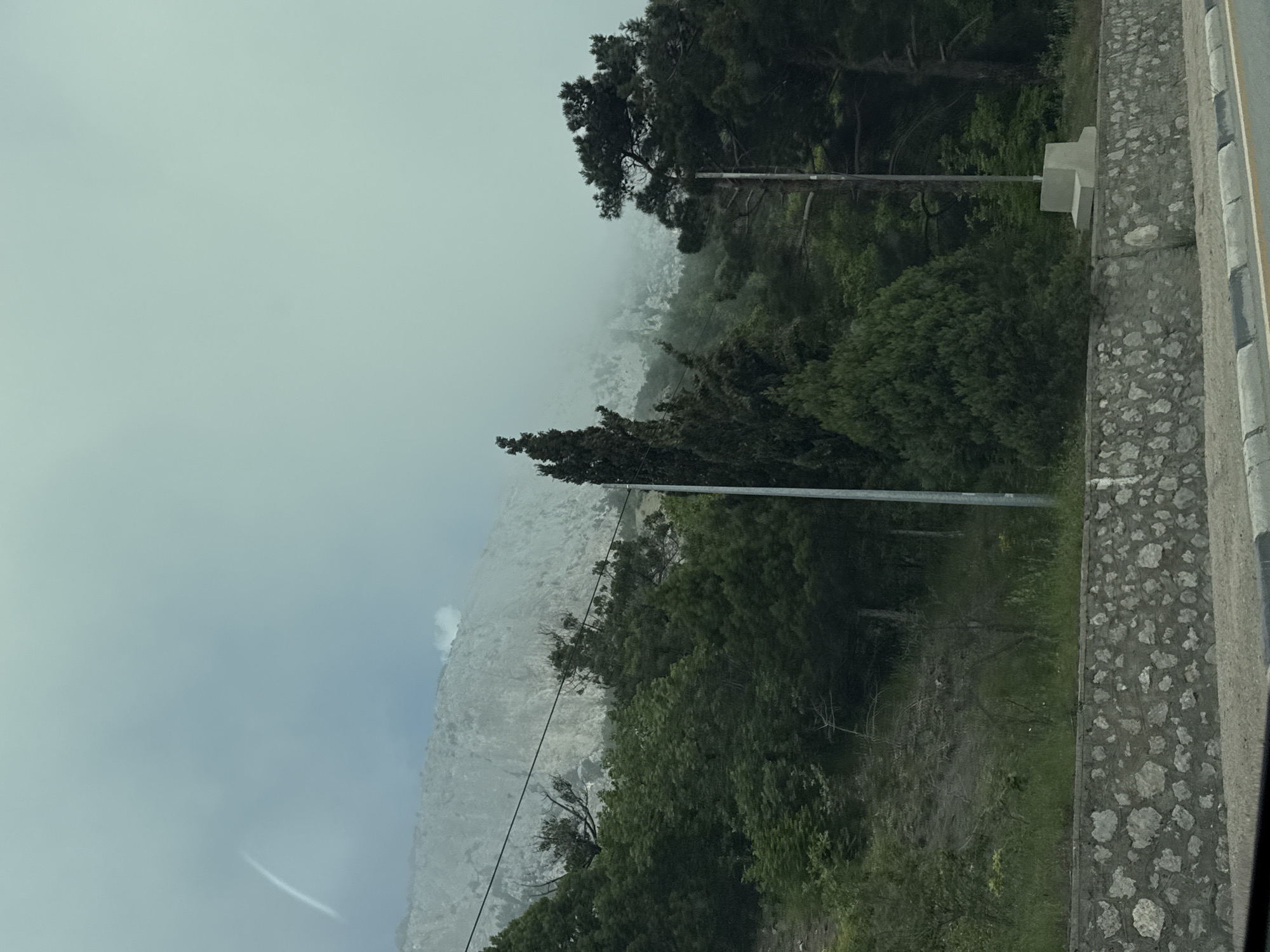 The image depicts a foggy landscape. In the foreground, there is a stone wall. Behind the wall, there are trees. In the background, there is a mountain covered in fog. The location is Unknown, Unknown.