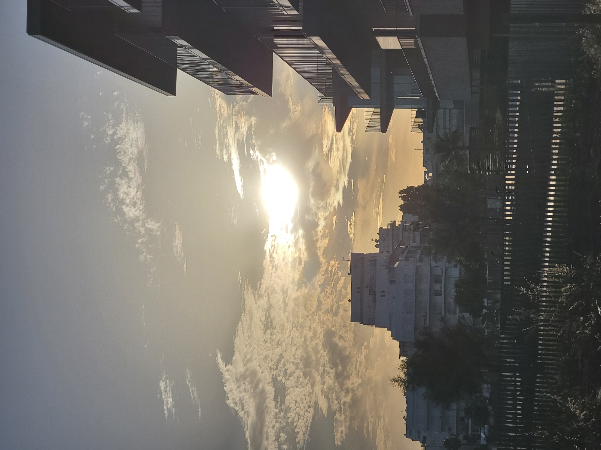 The image shows a sunset scene in Quarteira, Portugal, with modern buildings in the foreground and background. On the right, a multi-story building with balconies, designed in a minimalist style with dark grey or black railings, dominates the foreground. The sun is low in the sky, casting a warm, golden light that illuminates scattered clouds. In the background, to the left, a large, white, stepped building, characteristic of Quarteira's architecture, is visible. Between the foreground and background structures, a dark wooden fence runs horizontally across the lower part of the image, separating the viewer from a dimly lit foreground area with some vegetation. The sky is a gradient of pale blue and yellow, with the sun appearing as a bright, diffused orb. No people or animals are visible in the image.