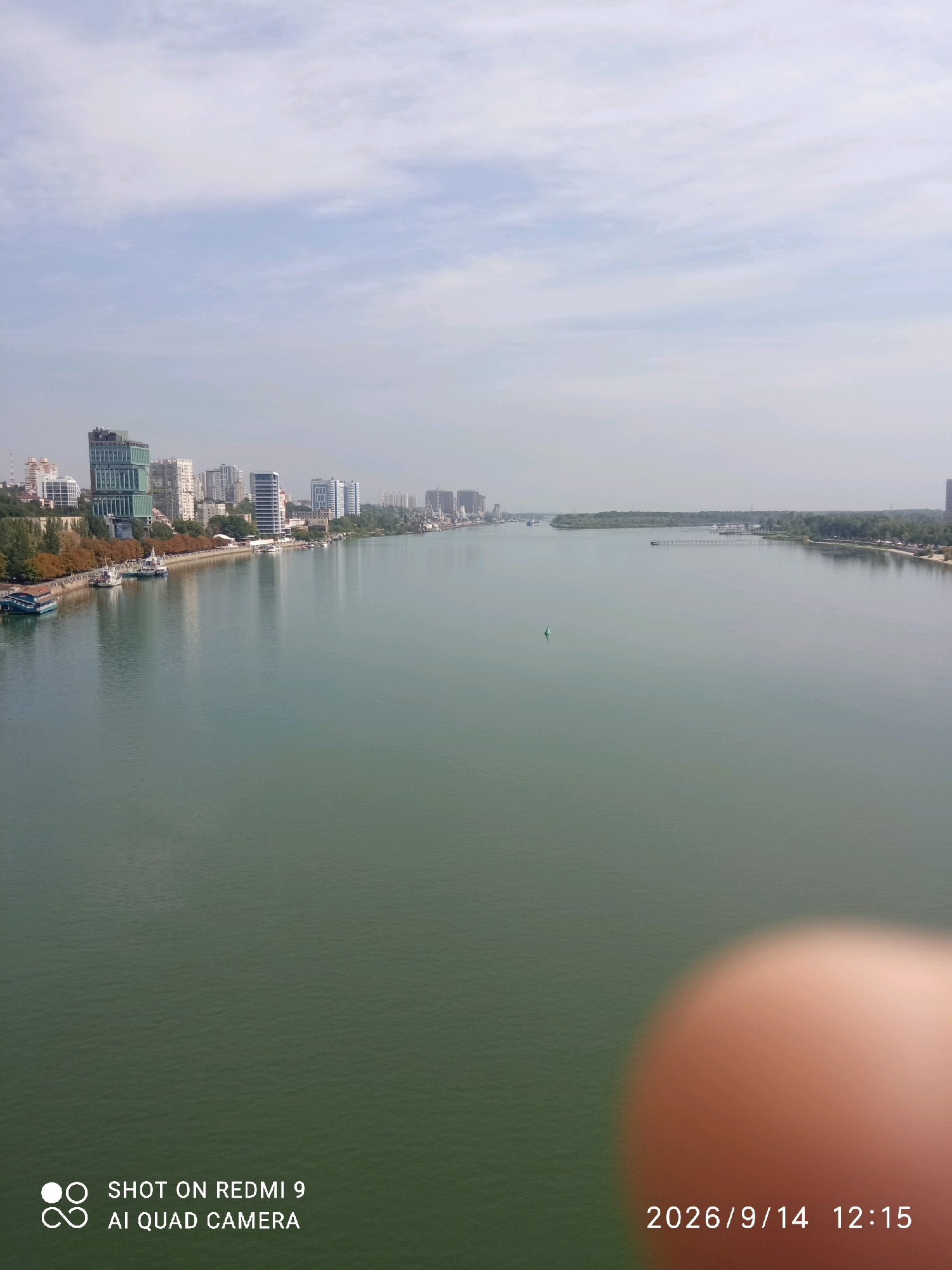 The image depicts a wide river with a city skyline on the left bank. The city features numerous modern high-rise buildings and older structures, interspersed with trees and a waterfront promenade. Several boats are docked along the shore. The river itself is calm and reflects the sky and buildings. In the distance, the river widens and is bordered by lush green trees on the right bank, with a sandy beach area visible. The sky is partly cloudy. In the foreground, a blurred, orangish circular object, likely a finger or thumb, obscures a portion of the lower right of the image. Text overlays indicate "SHOT ON REDMI 9 AI QUAD CAMERA" in the lower left and a date and time stamp "2026/9/14 12:15" in the lower right. This scene is consistent with the Rostov-on-Don embankment along the Don River.