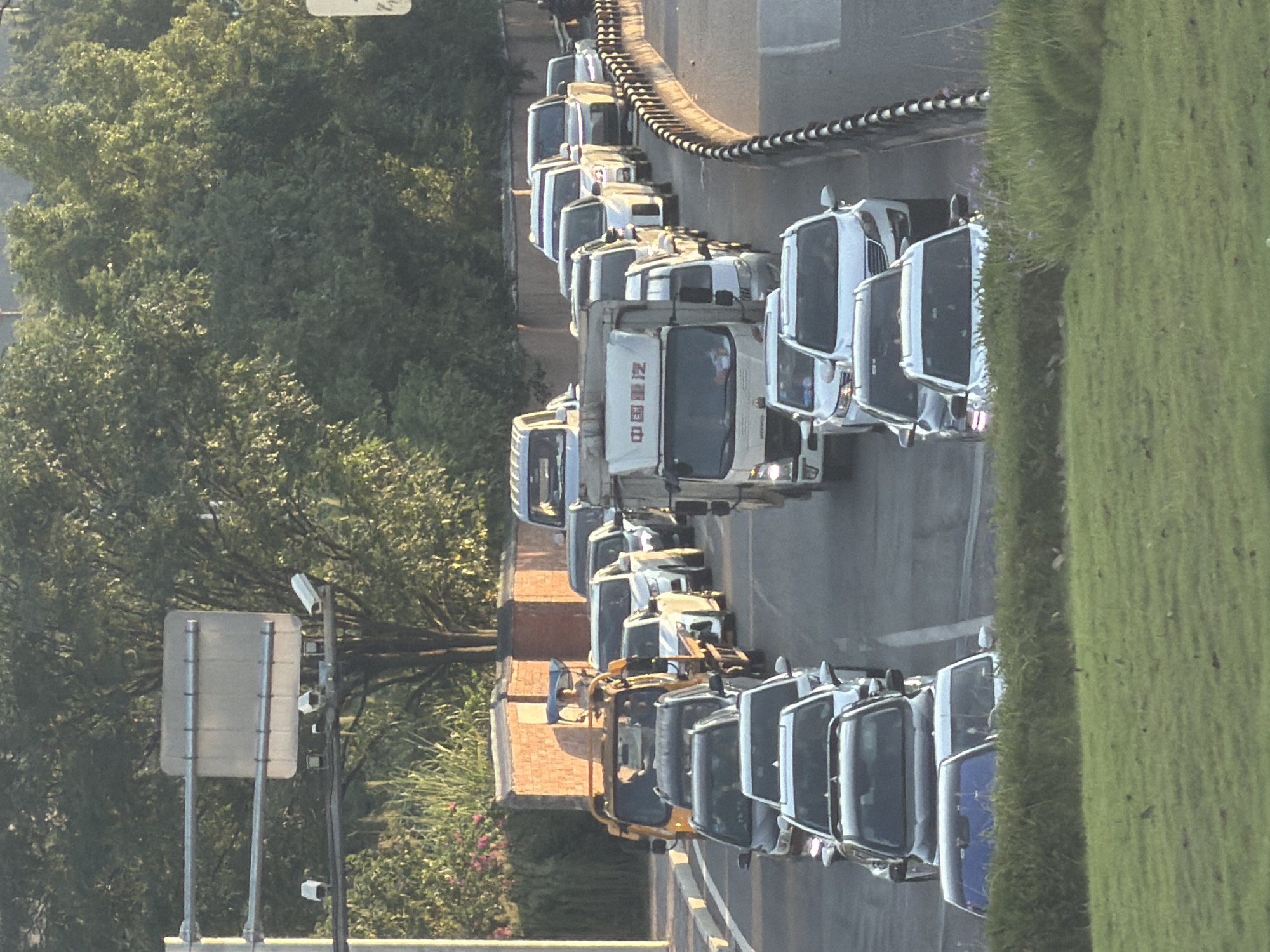 The image captures a traffic jam on a road in Jiangjin, China. The sun is setting, casting a warm glow on the scene, and the sky is clear. The road is lined with trees on the left and a brick wall with more trees behind it on the right. The traffic consists of various vehicles, including cars, trucks, and a tricycle. In the foreground, a grassy embankment is visible. On the left side of the road, there's a pole with traffic lights and surveillance cameras. The vehicles are stopped, indicating a standstill in traffic flow. The overall impression is one of a typical evening commute in a busy urban area.