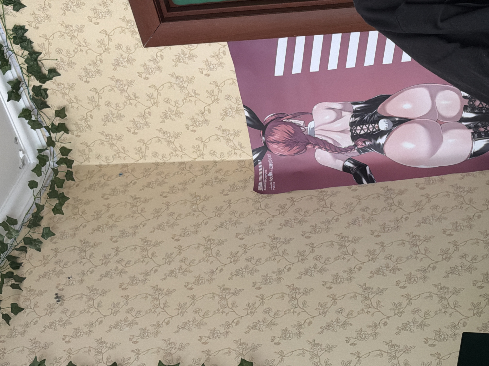 The image shows an interior corner of a room. The walls are covered with a light-colored floral wallpaper. A decorative string of green artificial ivy is draped along the top of the walls and ceiling. A large poster is leaning against the wall on the right side of the image. The poster depicts an anime character with bunny ears, wearing a black leotard and boots, with a braided red hairstyle. The character is shown from behind, with their back and buttocks prominently featured. Part of a dark-colored garment, possibly a jacket or hoodie, is visible in the lower right corner of the image, partially obscuring the poster. A wooden door frame is visible on the far right. The image was taken in Khimki, Russia.