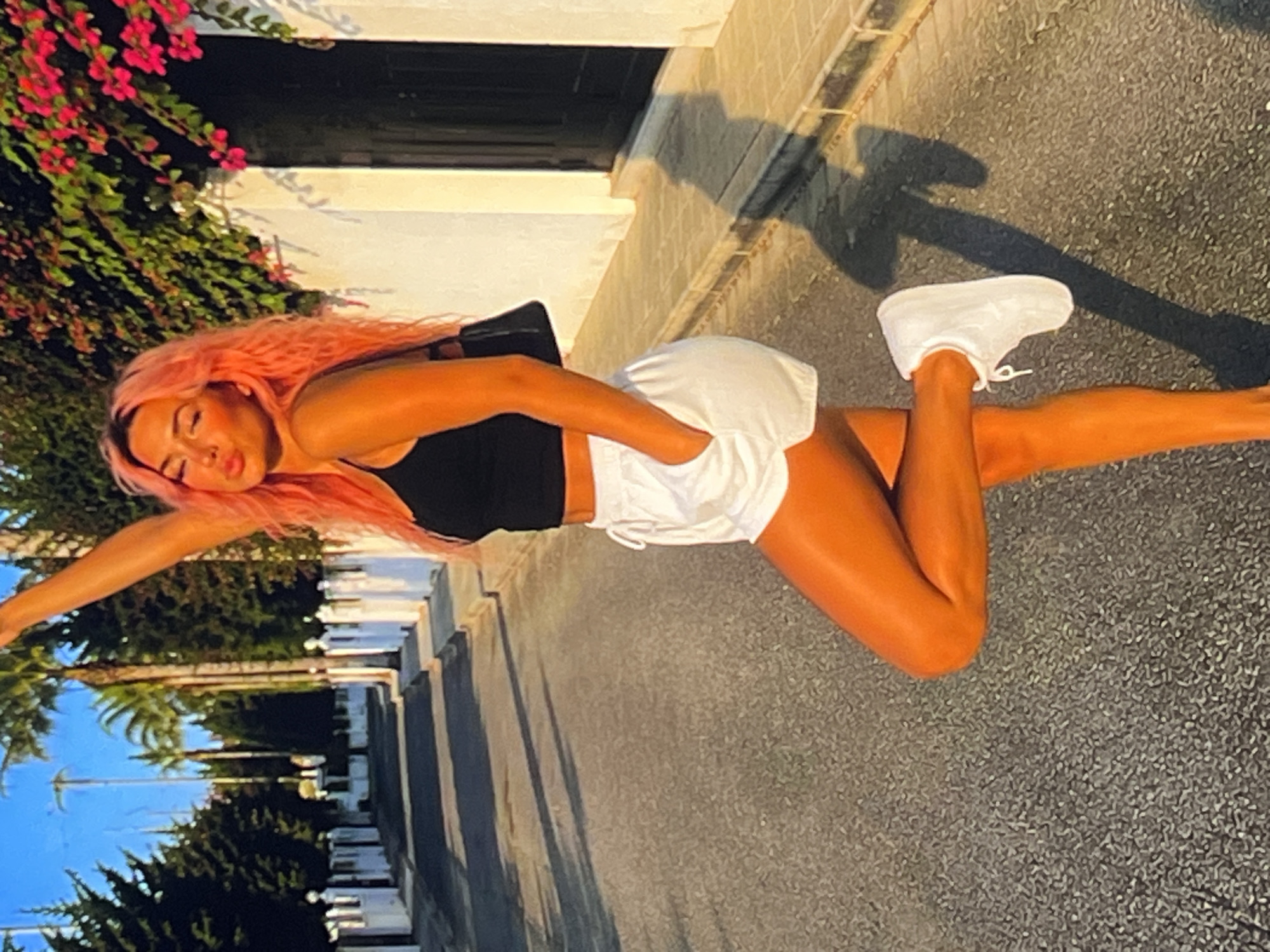 A young woman with bright pink hair is captured in a horizontal, outdoor shot, lying down with her arm extended and her head tilted back. She wears a black sports bra and white athletic shorts, accentuating her tanned skin. Her right leg is bent, with her white sneaker resting on the pavement, while her left leg is extended. The background suggests a sunny day, with long shadows cast on the asphalt path.
To the left of the woman, a vibrant green bush adorned with small pink flowers adds a touch of color to the scene, framing her upper body. Beyond the bush, a building with a white facade and a dark doorway is visible. The path is lined with trees, and a glimpse of a bright blue sky can be seen in the distance. The overall lighting and the long shadows indicate that the photograph was taken during the golden hour, likely late afternoon or early evening. The woman's relaxed pose and expression convey a sense of ease and enjoyment. There is no visible text in the image.