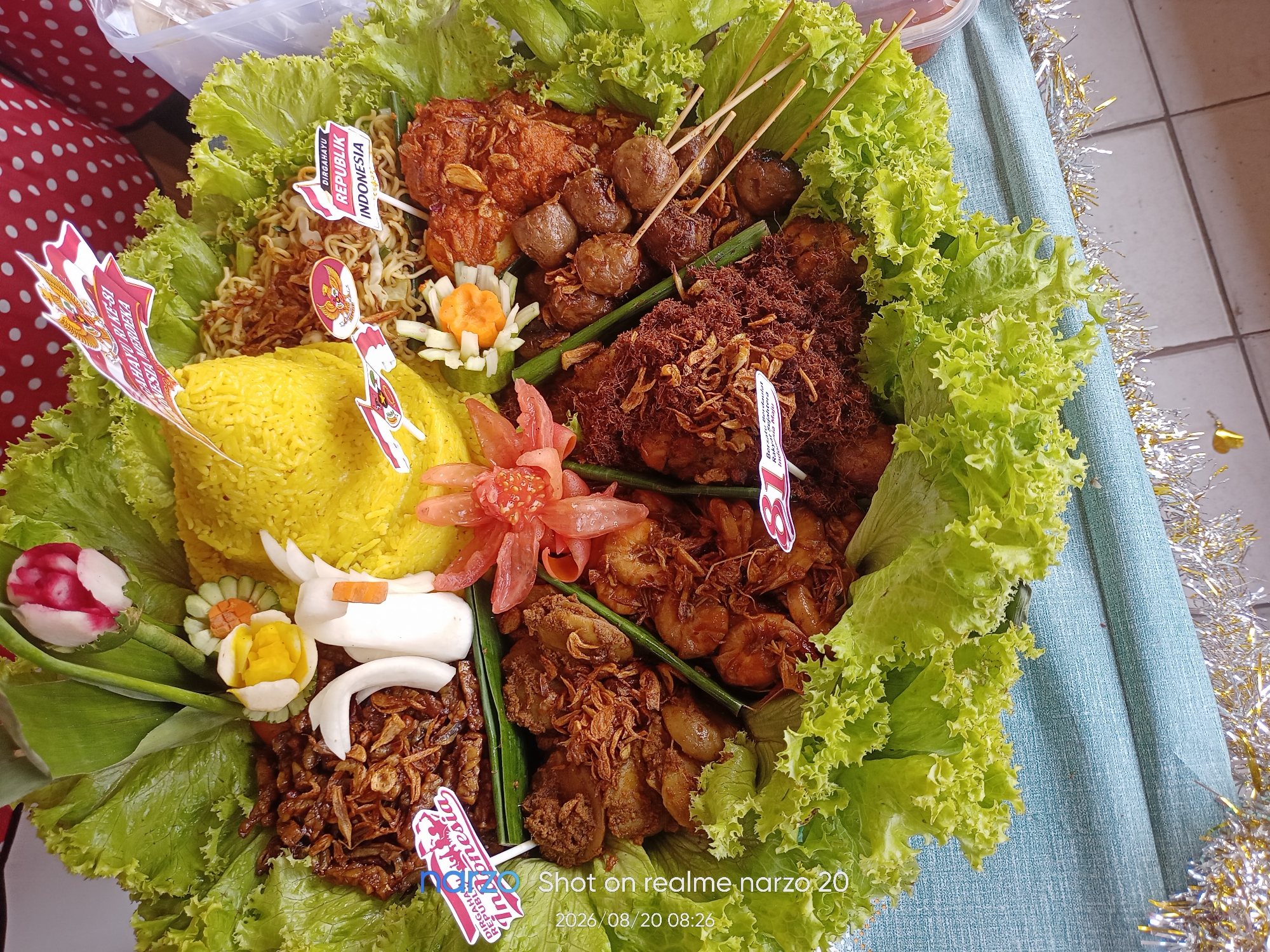 This is a high-angle, medium shot of a large, round platter of Indonesian food, likely a nasi tumpeng, artfully arranged and decorated. The platter is overflowing with various dishes, primarily savory. The base of the platter is lined with crisp, bright green lettuce leaves. In the center, a conical mound of vibrant yellow rice forms the core of the dish. Surrounding the rice are several distinct food items:
* On the left, there are mounds of fried noodles sprinkled with fried shallots.
* Central to the dish are several skewers of grilled meatballs.
* Adjacent to the meatballs are pieces of fried chicken covered in a reddish sauce and topped with fried shallots.
* Further into the platter, there are portions of what appears to be shredded or spiced chicken, also garnished with fried shallots.
* There are also several pieces of cooked shrimp.
* To the right of the yellow rice, there are decorative elements made from vegetables, including what looks like a carved radish or turnip and cucumber slices. There are also what appear to be pieces of boiled egg and some white, possibly radish, elements carved into a duck shape.
* A bright pink flower-like decoration, possibly made from chili or tomato, is placed near the center.
* Several small Indonesian flags with red and white colors and emblems are stuck into various parts of the food.
* The platter is situated on a light blue tablecloth that has a subtle texture.
* To the right of the platter, strands of silver and gold tinsel are visible, suggesting a festive occasion.
* In the bottom right corner, a watermark indicates the photo was "Shot on realme narzo 20" and dated "2026/08/20 08:26".
The food appears to be prepared and presented for a celebration or event. The location is identified as Karawang, Indonesia.