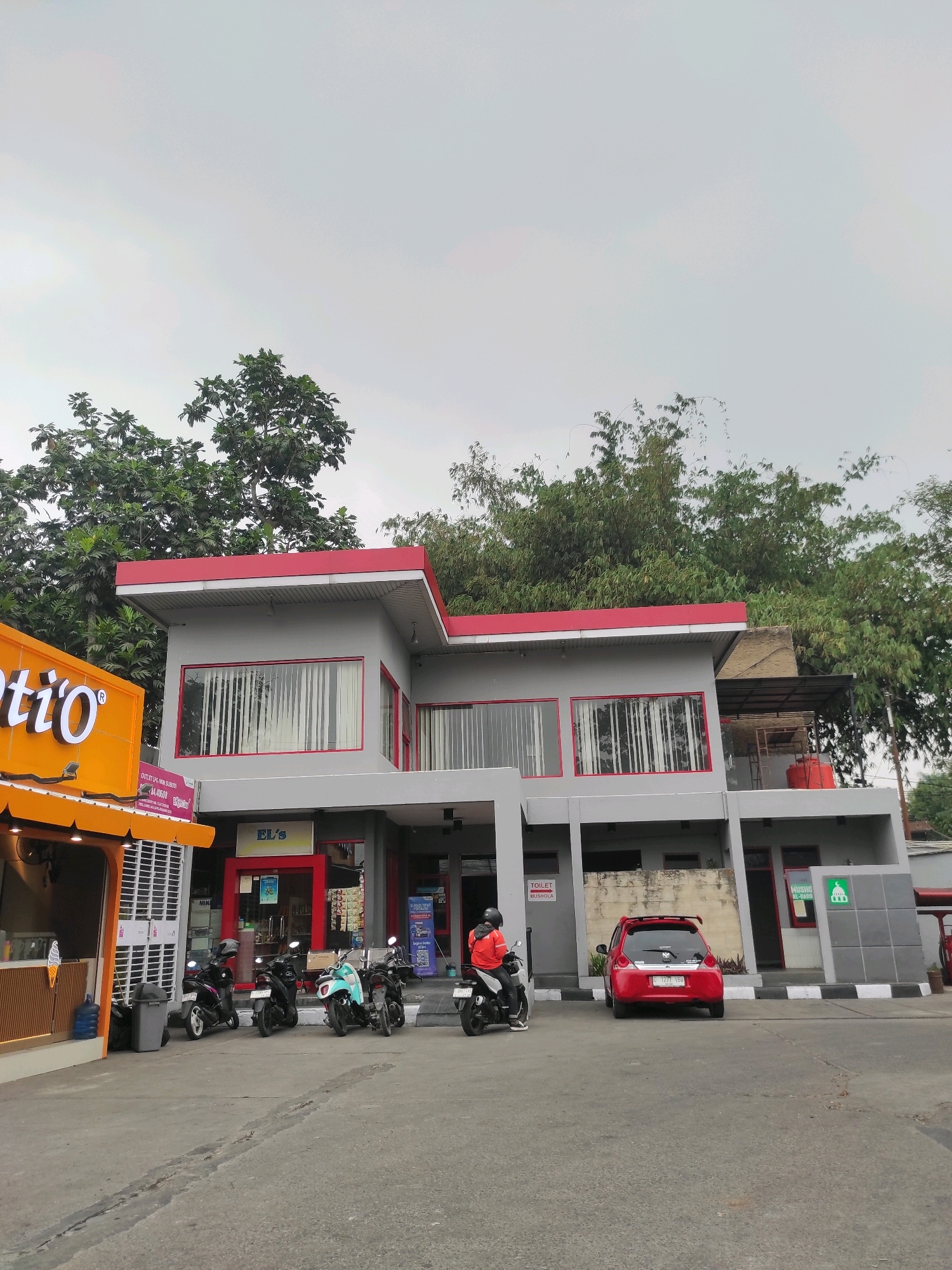 A red car is parked in front of a modern, gray building with red trim. The building appears to be a commercial establishment, possibly a gas station or convenience store, with signage including "EL's" and "MUSHOLLA AL-BADR". Several motorcycles are parked in front of the building, and one person is seated on a motorcycle wearing an orange jacket. To the left, a bright orange building with a sign reading "atio" is visible. Trees and bamboo are in the background, under an overcast sky. The scene is set on a paved area, likely a parking lot or road.