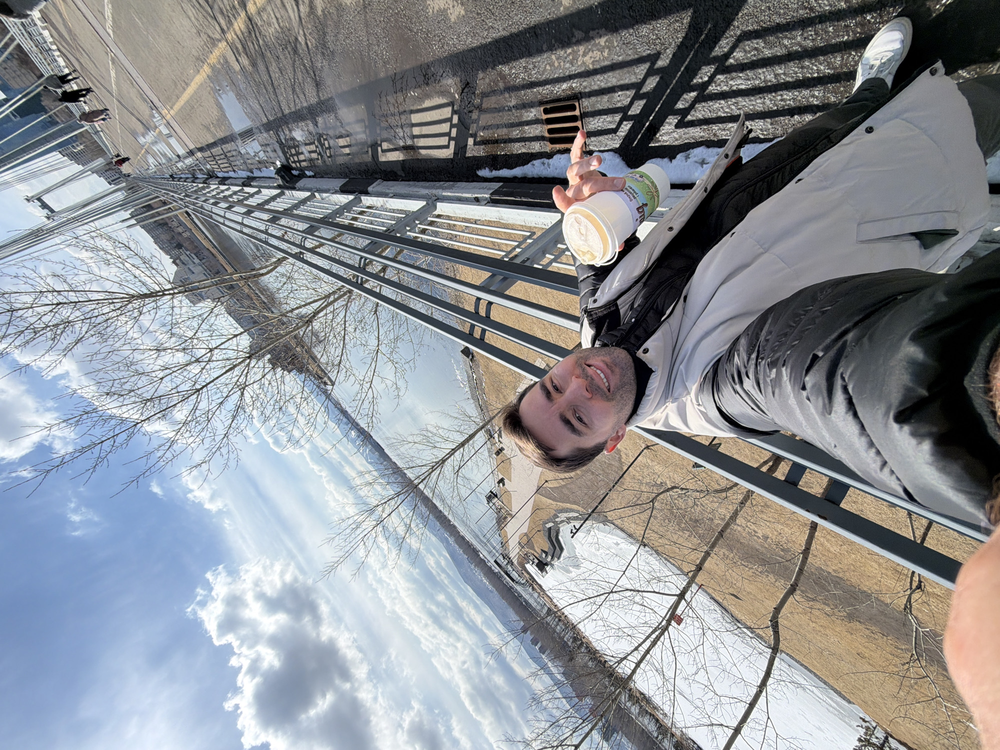 A selfie-style photograph captures a young man outdoors on what appears to be a bridge or elevated walkway in Krasnoyarsk, Russia. The image is taken from a slightly low angle, looking upwards and outwards.
The main subject, a man with short dark hair and a light beard, smiles faintly towards the camera. He is dressed warmly in a light-colored (likely off-white or light grey) long winter coat over dark clothing and dark pants, suggesting cool weather. In his right hand, he holds a white cup with a green label that displays visible Cyrillic text, including "Биологически активная добавка" (Biologically active additive) and "Донат...".
The setting is an elevated structure with a metal railing to the man's left. The ground is an asphalt surface with a yellow line, and patches of melting snow are visible alongside the curb and a storm drain. Long, distinct shadows cast by the railing indicate bright sunlight. In the distance on the walkway, other pedestrians and pigeons are visible.
Beyond the railing, the scene opens up to a vast, partially frozen body of water, likely the Yenisei River. Bare tree branches frame the left and right sides of the image, characteristic of late winter or early spring. Across the river, an urban landscape unfolds with various buildings and structures, under a clear blue sky with scattered white clouds. The bright daylight and long shadows suggest it is either mid-morning or late afternoon. The man's smile conveys a pleasant mood while enjoying the scenic view.