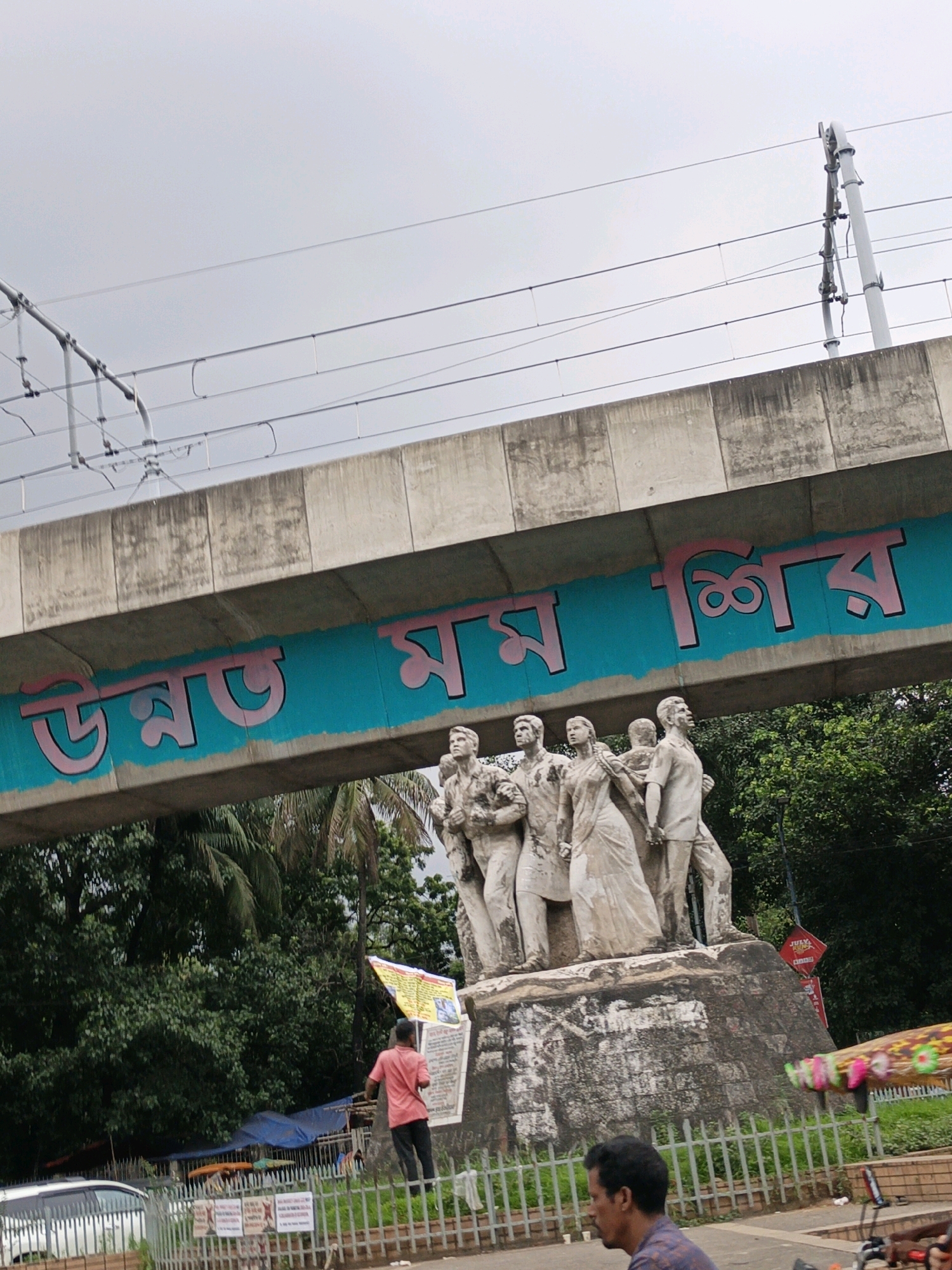 The image shows a statue of five people standing on a stone pedestal. The statue is located in Dhaka, Bangladesh. The statue is in front of a bridge with Bengali writing on it. The writing on the bridge says "উন্মোচন". The sky is cloudy. There are trees in the background. A person in a pink shirt is standing near the statue, looking at a banner. There is a fence in front of the statue. A car is parked on the left side of the image.