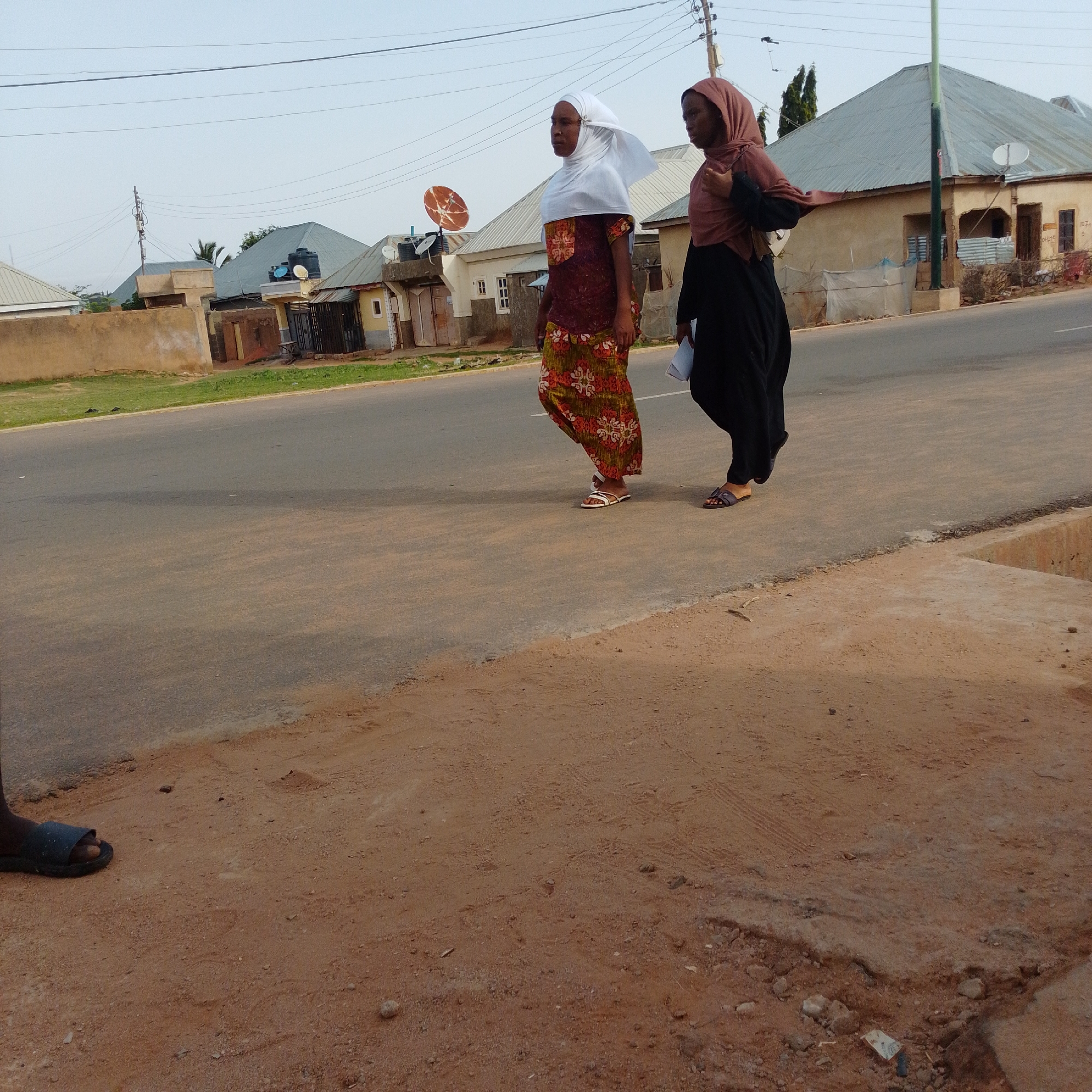 In the city of Gombe, Nigeria, two individuals are captured in a street scene. One person, wearing a white headscarf and a colorful patterned dress, walks on the asphalt road. Beside them, another person, dressed in a dark garment and a reddish-brown headscarf, also walks on the road. In the background, several buildings with corrugated metal roofs are visible, along with utility poles and overhead wires, typical of an urban setting. The ground in the foreground is a reddish-brown dirt. The lighting suggests it is daytime.