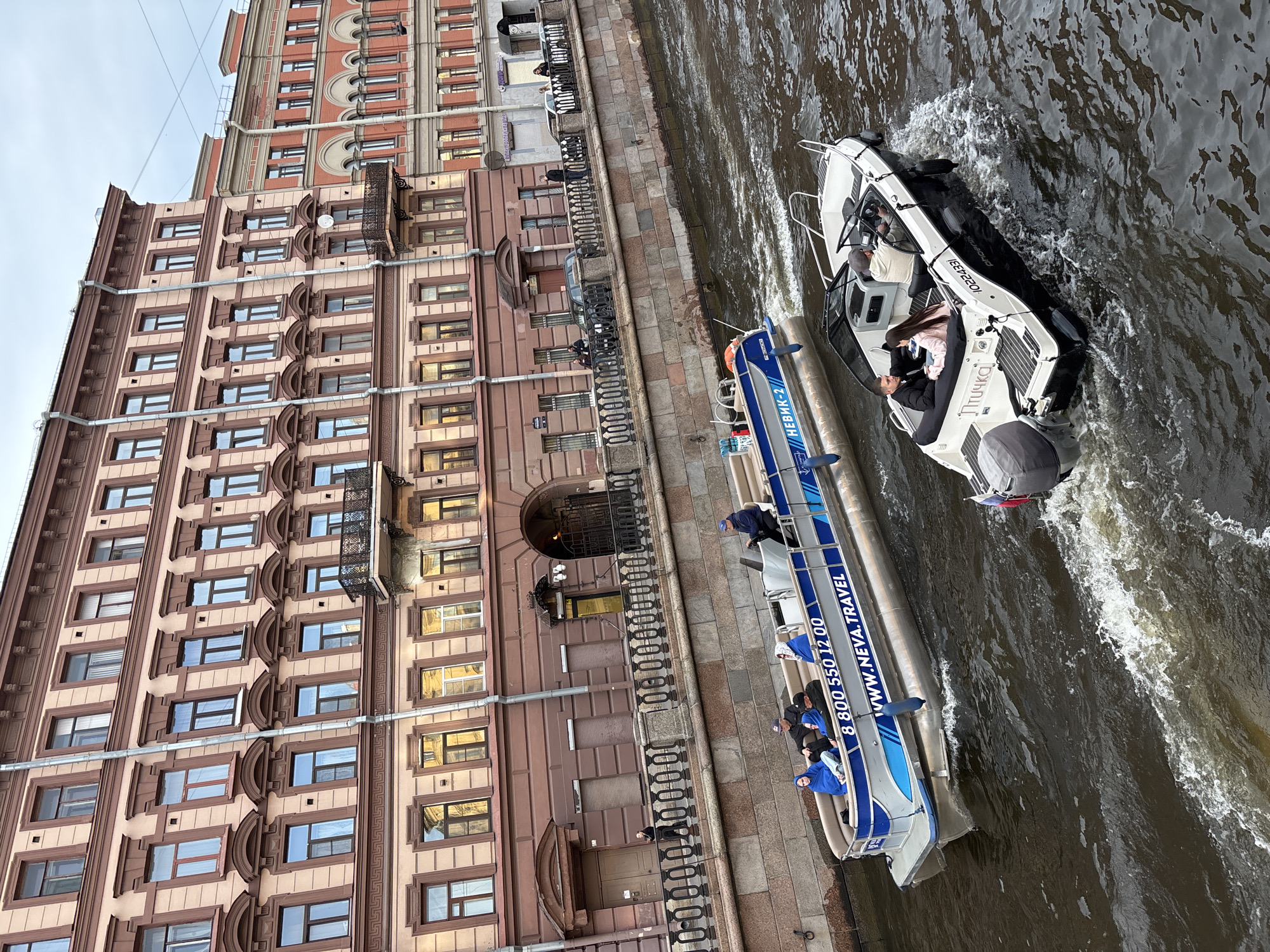 This image captures a vibrant canal scene in Saint Petersburg, Russia, viewed from an elevated, slightly tilted perspective. A grand, multi-story historic building with elaborate brown and beige facades, featuring classical architectural elements such as arched windows, pilasters, and decorative cornices, lines the stone embankment. Its numerous windows reflect the overcast daylight.
Two boats are navigating the dark, choppy water of the canal. To the left, a long, blue and white tour boat is prominently labeled "НЕВИК-2" (NEVIK-2), "WWW.NEVA.TRAVEL", and displays a phone number: "8 800 550 12 00". Several passengers, some wearing blue jackets, are seated in the boat, with a person in a blue cap standing near the bow, indicating a guided tour on the Neva River or its associated canals.
Alongside it, to the right, a smaller, private motorboat, predominantly white and black, is also in motion, creating a wake. This boat bears the name "Птичка" (Ptichka, meaning "little bird"), the brand "phoenix", and a registration number "10224331". At least two people are visible onboard: a man at the helm and a woman (and possibly another person) seated in the passenger area, enjoying a recreational ride.
The scene depicts a casual daytime activity under an overcast sky, showcasing the architectural beauty of Saint Petersburg from its waterways. The visible text "NEVA.TRAVEL" strongly ties the location to the Neva River, a central feature of the city.