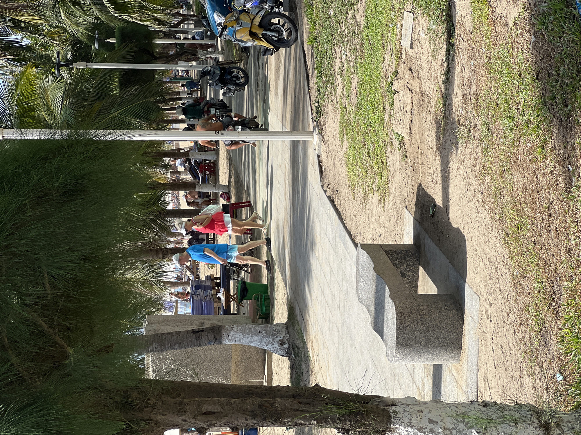 In Nha Trang, Vietnam, a paved walkway lines a sandy beach area shaded by palm trees and other foliage. A man in a blue shirt and shorts, and a woman in a red top and white skirt walk away from the camera on the walkway. Several motorbikes are parked along the right side of the walkway, with a yellow scooter prominently in the foreground. In the background, beach chairs and umbrellas are visible, indicating a public beach setting. A distinctive stone bench is situated on the sandy area to the left of the walkway.