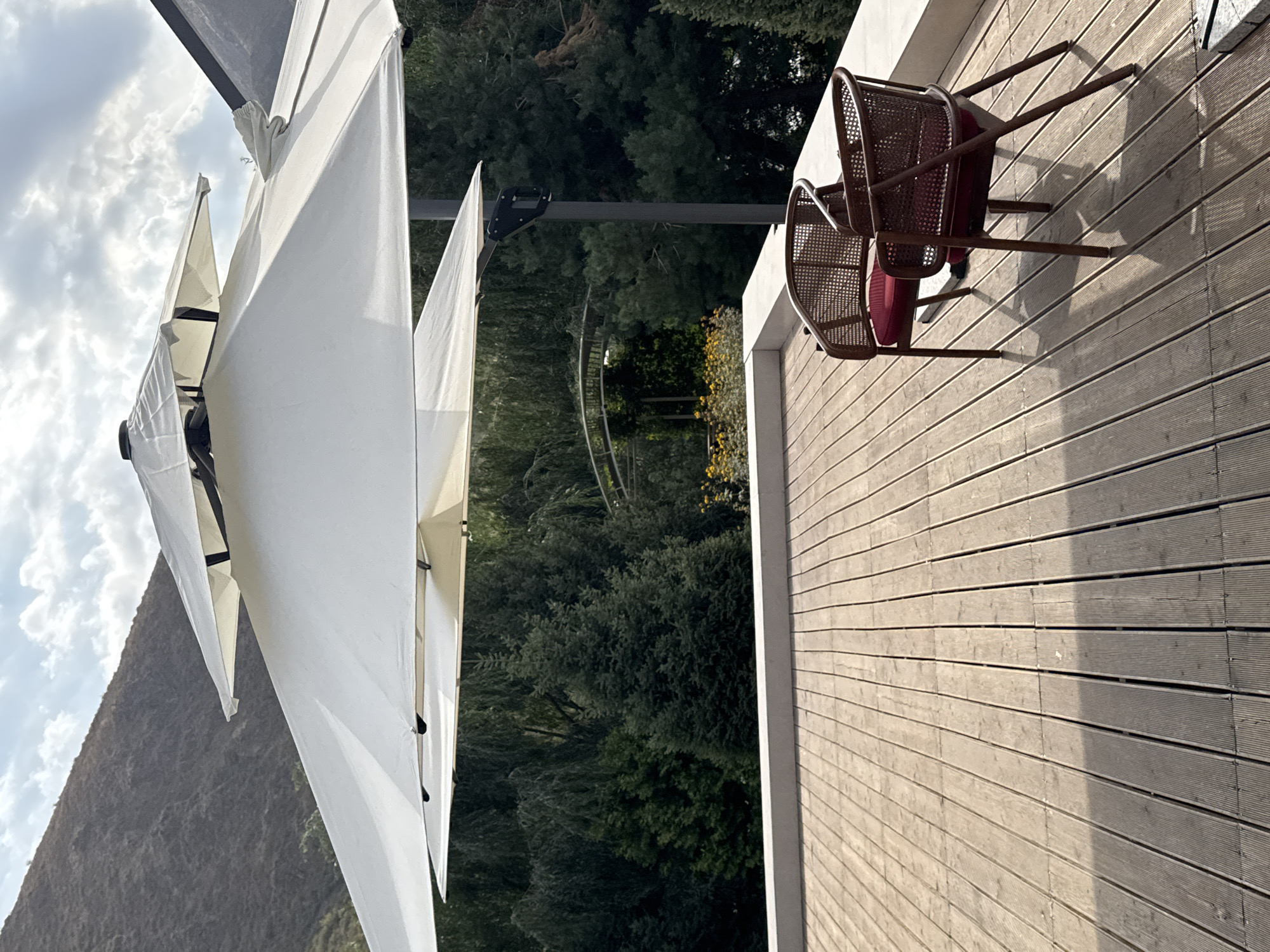 This is a vertical, eye-level shot of a wooden deck in Koy Tash, Kyrgyzstan, featuring two empty wicker chairs with red cushions. The chairs are positioned in the foreground, with their shadows cast on the wooden planks. In the background, a large, tiered white umbrella provides shade over the deck. Beyond the umbrella, a dense forest of dark green trees creates a lush backdrop, with hints of a mountainous landscape visible in the upper left corner. The sky is partly cloudy, with sunlight breaking through. The overall scene suggests an outdoor seating area in a natural, possibly mountainous, setting.
