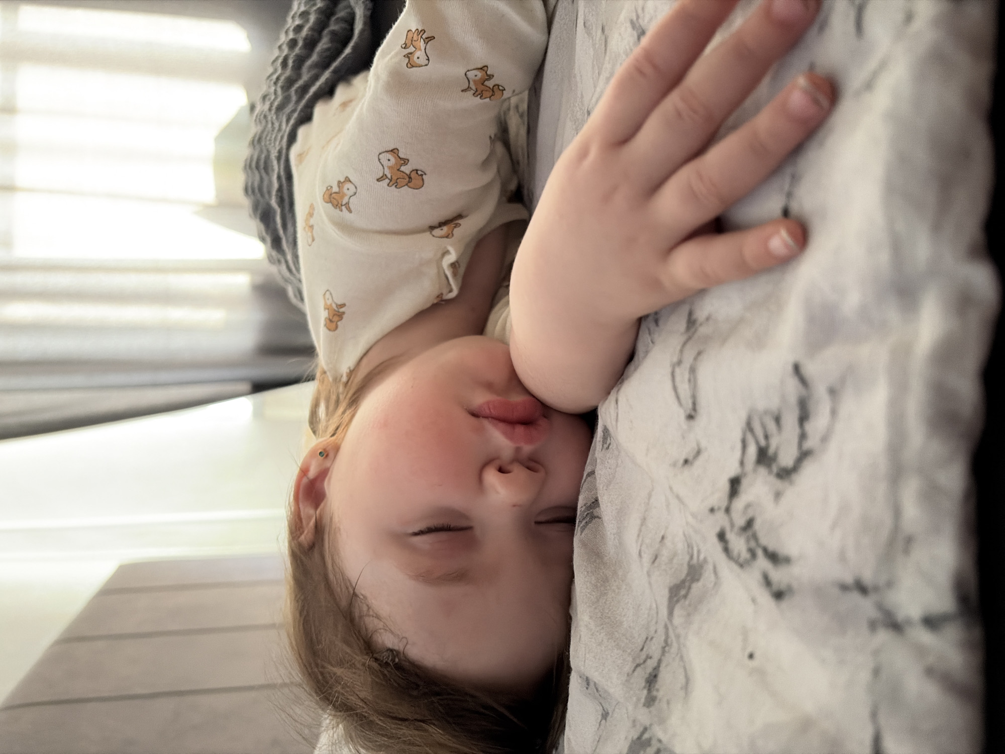 A young child with fair skin and light brown hair sleeps on a bed. The child is wearing a light-colored long-sleeved shirt adorned with fox patterns. Their hand rests on a white and gray patterned pillow. A grey knit blanket is draped above the child. The location is not evident from the image.