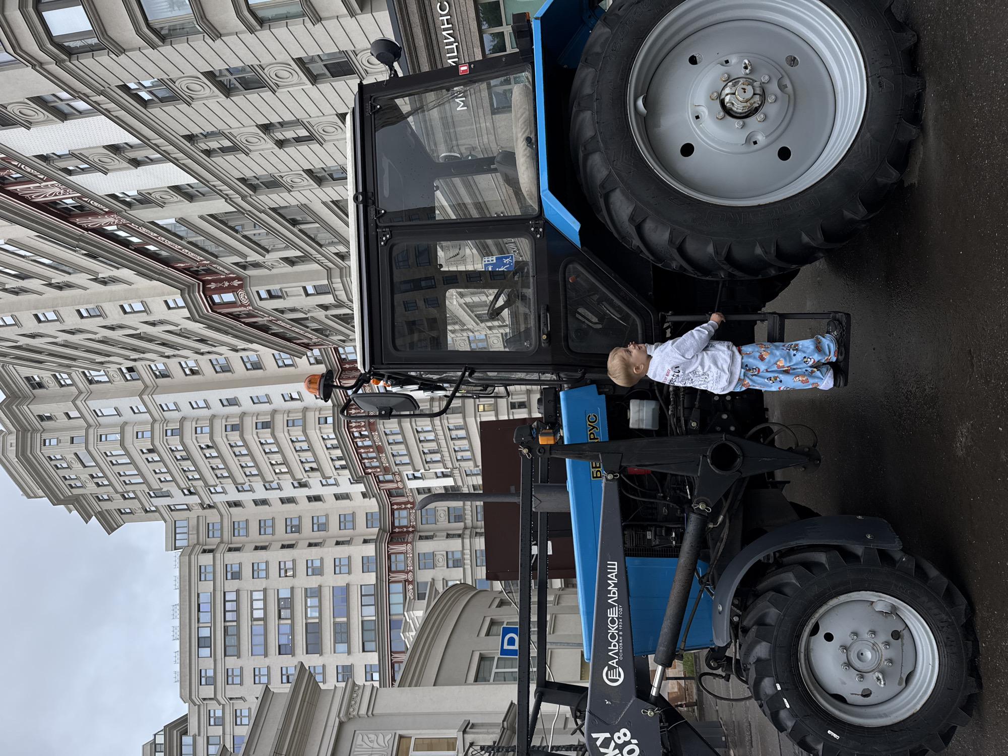 The image depicts a young boy in a light gray sweatshirt and pajama pants standing on the steps of a large blue tractor. The tractor has a black cab with tinted windows and a large rear tire. The tractor is parked on a wet paved surface, likely a street or square, with the rear of a large building visible in the background. The building is an ornate, multi-story structure with numerous windows and decorative architectural elements. The sky is overcast and gray. Text on the tractor's loader arm reads "ЕЛЪСКОЕАЛЬМАШ" and "ОСНОВАН В 1934 ГОДУ". The text on the blue body of the tractor appears to be in Russian. The overall scene suggests an urban setting, likely a public event or display in Moscow, Russia, given the architectural style and the presence of Cyrillic text.
