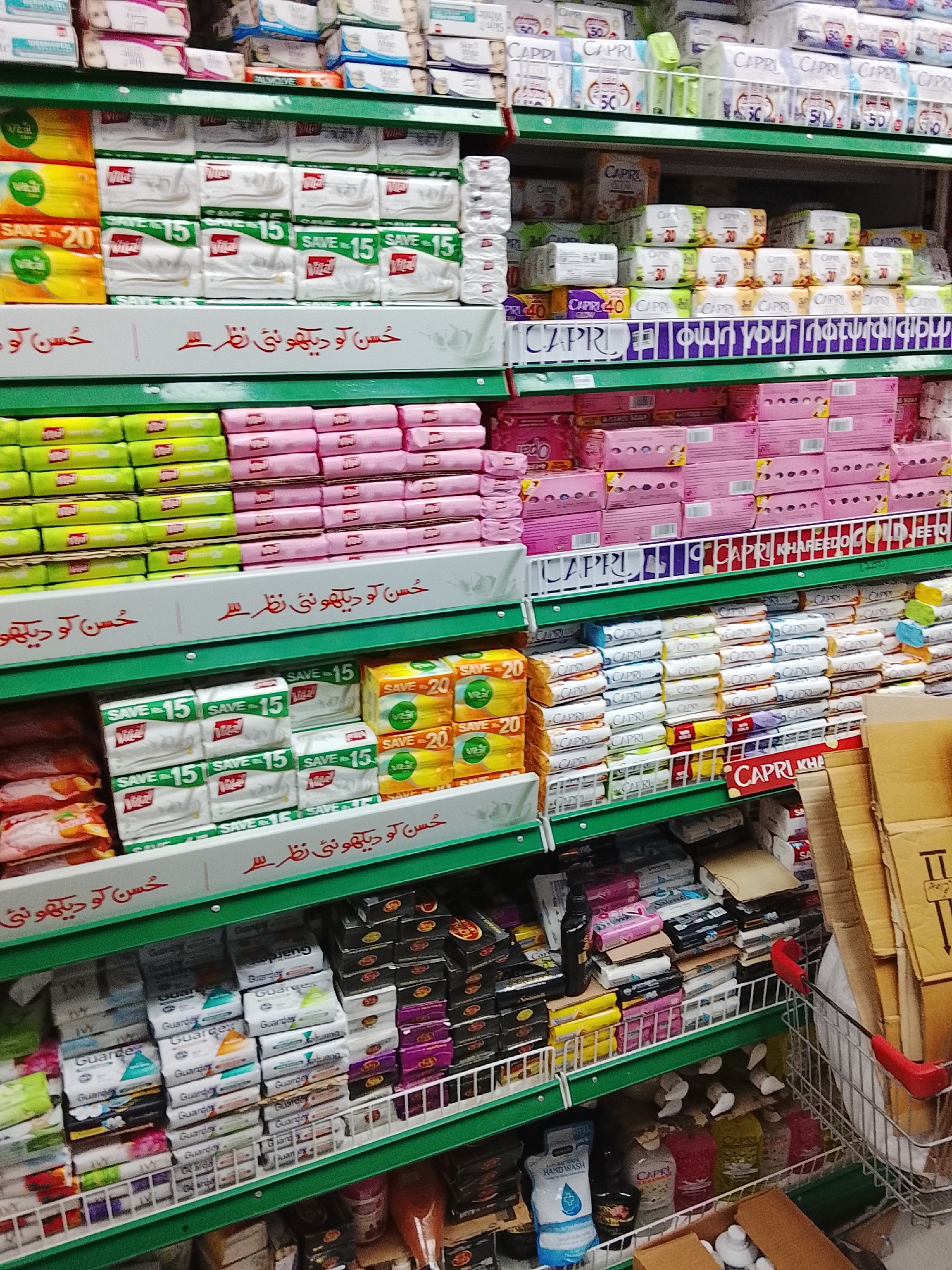 The image depicts a shelf in a retail store, possibly a supermarket, stocked with various hygiene and personal care products. The shelf is divided into multiple levels. The top shelf contains items like Capri soap, and other sanitary products. The middle shelves have Vita soap, and other brands. The bottom shelves house products like Guardian soaps and hand wash. The shelves are labeled in Urdu. A shopping cart filled with cardboard boxes is visible on the right. The location is likely Gulgasht Colony, Pakistan, based on the language used on the shelves.