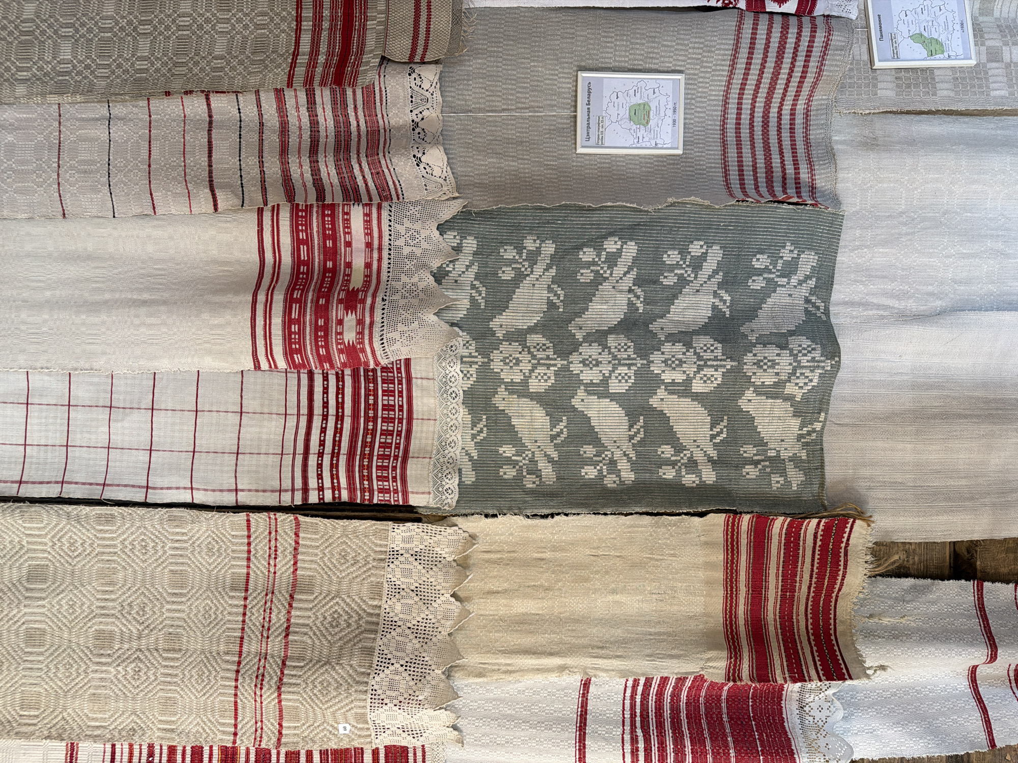 The image displays a collection of traditional Belarusian textiles, likely from the region of Літвенскі сельскі Савет, Belarus. The textiles are arranged vertically and appear to be hung on a wall, possibly in a museum or exhibition. There are several distinct patterns and colors: one textile features a geometric diamond pattern in beige and red stripes, another has a red and white checkered pattern with red stripes at the bottom, and a third has a green background with a white bird and floral motif. There are also plain white and cream-colored textiles with subtle woven patterns and red stripes. Two framed informational signs are visible on the wall. One sign, positioned on a grey textile with red stripes, displays a map of "Central Belarus" and text in Cyrillic, with a date range of 1930-1950. Another sign, visible lower down on a patterned textile, is also a map with Cyrillic text and the date 1930. The overall impression is one of cultural heritage and craftsmanship.