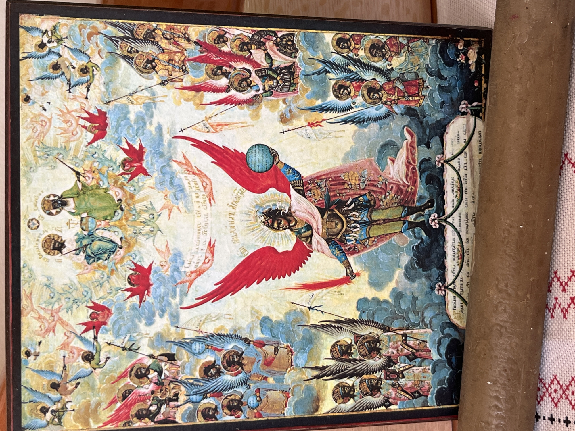 An icon depicts a central winged figure holding a globe, surrounded by numerous smaller winged figures and celestial beings. The central figure has large red wings and wears ornate armor and a pink cloak. They hold a blue globe in their left hand and a sword in their right. Above and to the right of the central figure, two larger figures, likely divine beings, are seated on thrones, one in green and the other in blue, with halos. Surrounding these figures are many smaller, winged beings, some with halos, some carrying scrolls or weapons. Some of the smaller figures are depicted as cherubic, while others appear more human-like. The background is a mix of celestial blue and clouds, with streaks of gold and pink suggesting divine light or energy. At the bottom of the icon, there is text inscribed in an unfamiliar script, along with floral motifs. The overall style is typical of Eastern Orthodox iconography, with flattened perspectives and symbolic representation. The icon is framed in a dark border. The image is taken from a slightly angled, head-on perspective, with a brown, cylindrical object in the foreground obscuring the bottom portion of the icon and a patterned textile beneath it. The location is described as Unknown, Unknown.