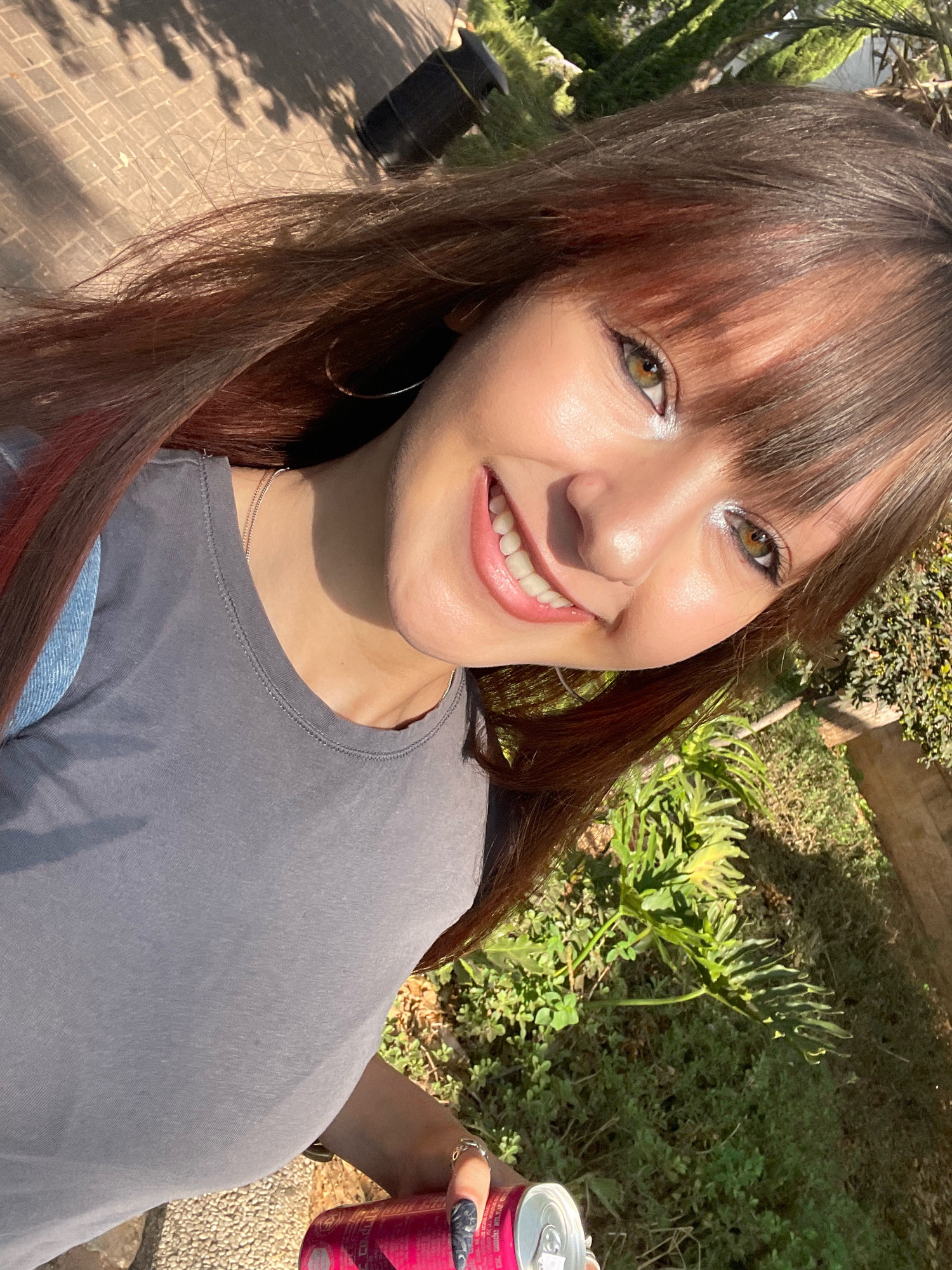 A young woman with long, brown hair and bangs smiles at the camera, holding a pink can in her right hand. She is wearing a grey t-shirt and a silver necklace. The background shows a paved area with shadows cast by trees, green foliage, and a black cylindrical object. The lighting suggests it is daytime, with sunlight casting strong shadows. The image appears to be taken outdoors, possibly in a park or urban green space.