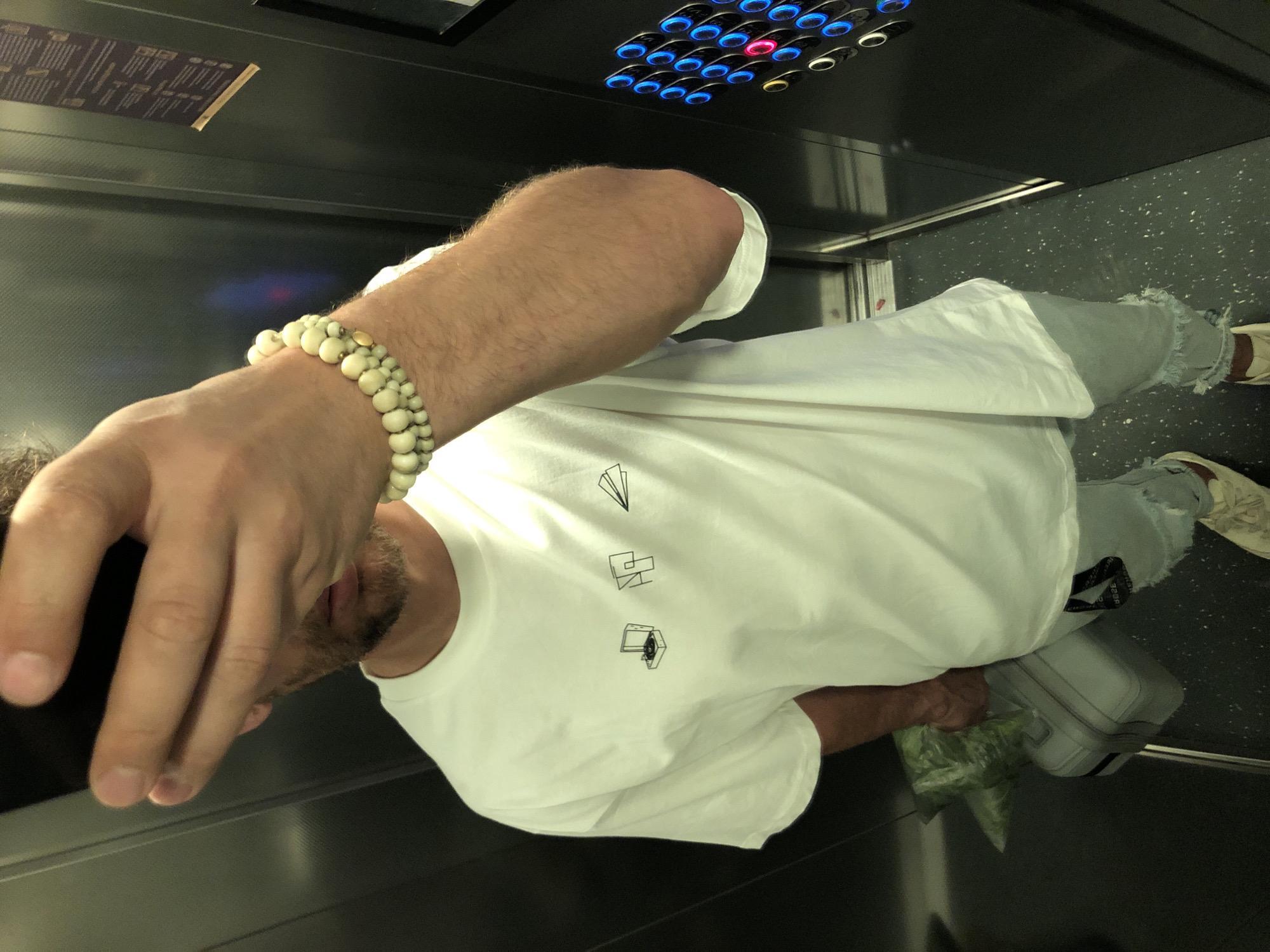 An adult male is observed inside an elevator in Saint Petersburg, Russia, taking a photograph from an elevated perspective, likely a selfie. He is wearing a loose-fitting white t-shirt adorned with three black line-art graphics: a record player, an abstract geometric shape, and a paper airplane. His lower attire consists of light blue, distressed jeans with frayed cuffs and light-colored sneakers featuring a maroon or brown side detail. His right arm is visible, adorned with a multi-strand bracelet composed of numerous spherical, cream-colored beads, accented by a single small gold-colored bead. His left hand holds a dark-colored smartphone, partially obscuring his face, which shows stubble. With his left hand, he also holds the handle of a light grey hardshell suitcase, on top of which rests a translucent green mesh bag, possibly containing produce. The elevator features dark, possibly metallic, wall panels and a dark speckled floor. On the right wall, there is a control panel with multiple circular buttons, predominantly illuminated in blue, with one button illuminated in red and another in yellow. Above the control panel, a brown rectangular notice or sign is affixed to the wall.