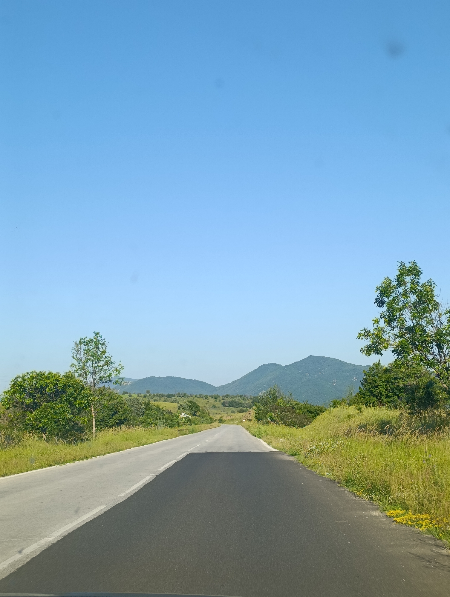 The image depicts a paved road receding into a landscape of rolling hills and mountains under a clear blue sky. The road is divided by a white line, with one lane appearing lighter in color, possibly due to recent resurfacing or a different material. Lush green grass and scattered trees line both sides of the road. In the distance, several green, tree-covered hills and mountains are visible. The scene appears to be captured from a vehicle, as suggested by the perspective and the lower edge of what could be a dashboard or windshield. The location is identified as Komuniga, Bulgaria.