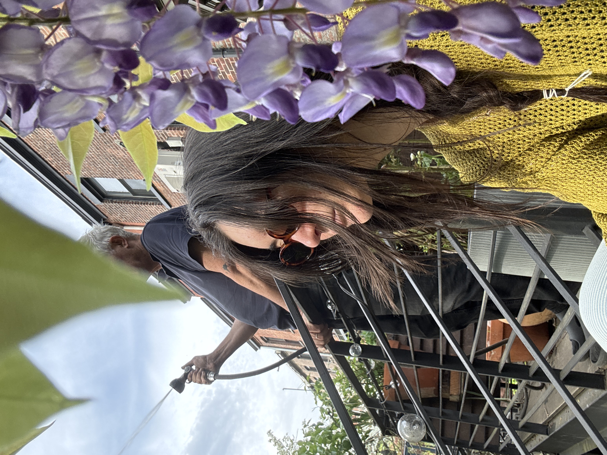 The photograph captures a scene outdoors, seemingly on a terrace or balcony, potentially within the city of New York, United States of America. The perspective is unique, as the photographer appears to be positioned above the subjects, looking down at an angle.
A woman with long, dark hair, wearing sunglasses, is positioned in the upper portion of the frame. She is wearing a yellow knit top. Above her head are purple flowers.
Below her, a man is seen holding a hose, from which water is being sprayed. The man is older, with gray hair. He is wearing a dark-colored t-shirt.
The background consists of an iron staircase or railing, suggesting a multi-level outdoor space. Additionally, a view of surrounding buildings and a partly cloudy sky are visible, indicating an urban environment.