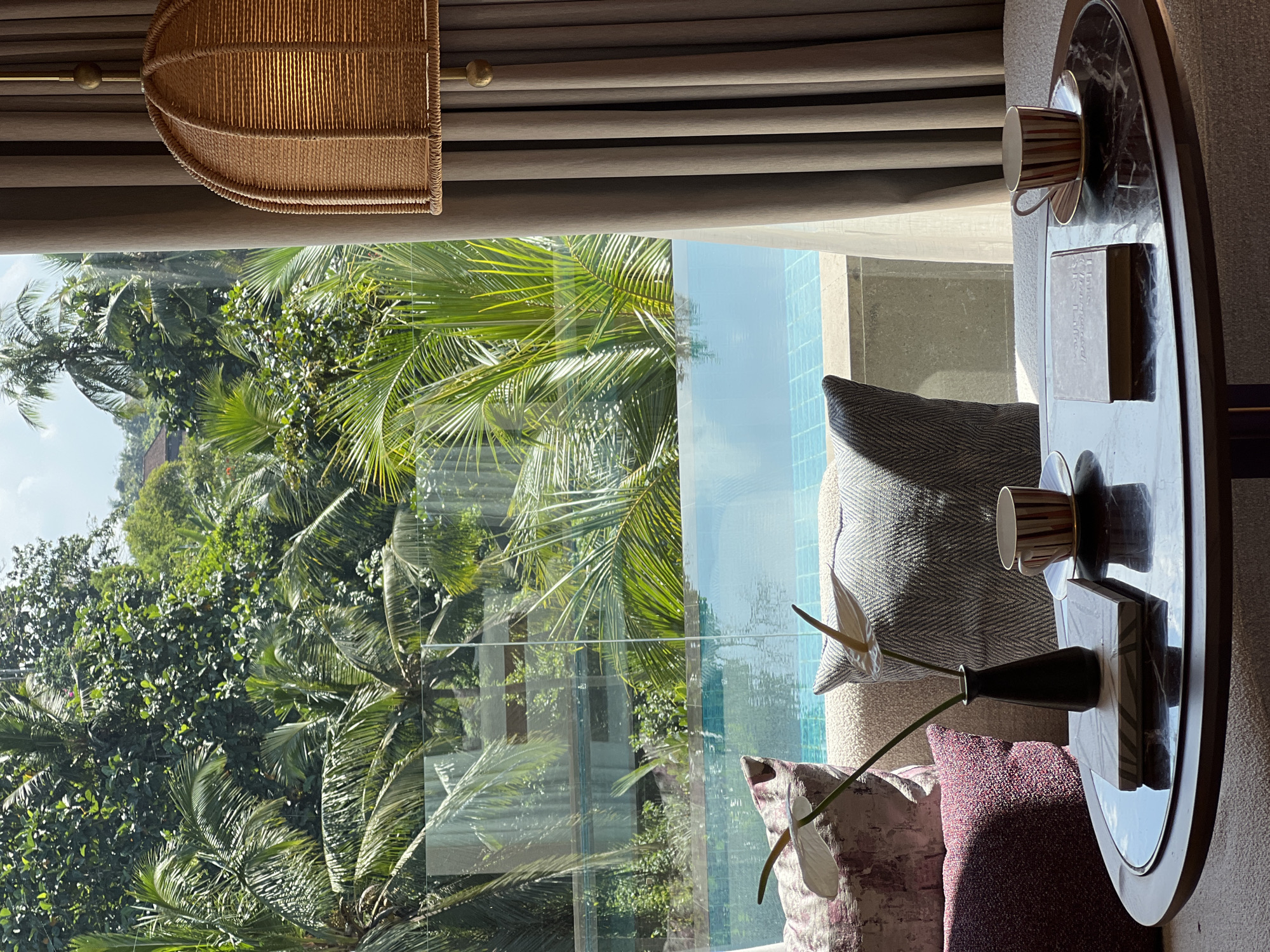 An interior shot from a room in Ubud District, Indonesia, shows a glimpse of lush tropical greenery and a swimming pool through a large window. In the foreground, a round, dark marble-topped table holds two coffee cups and saucers, a book, and a small black vase with two white flowers. The cups have a striped pattern. Behind the table, a sofa is adorned with several decorative pillows, one with a floral pattern and others with textured designs. To the right, a beige curtain is partially drawn, revealing a woven pendant lamp with a warm glow. The window offers a view of palm trees and dense vegetation, with the edge of an infinity pool visible below. The water of the pool reflects the sky and surrounding foliage. A building with a red roof is partially visible in the distance amidst the trees.